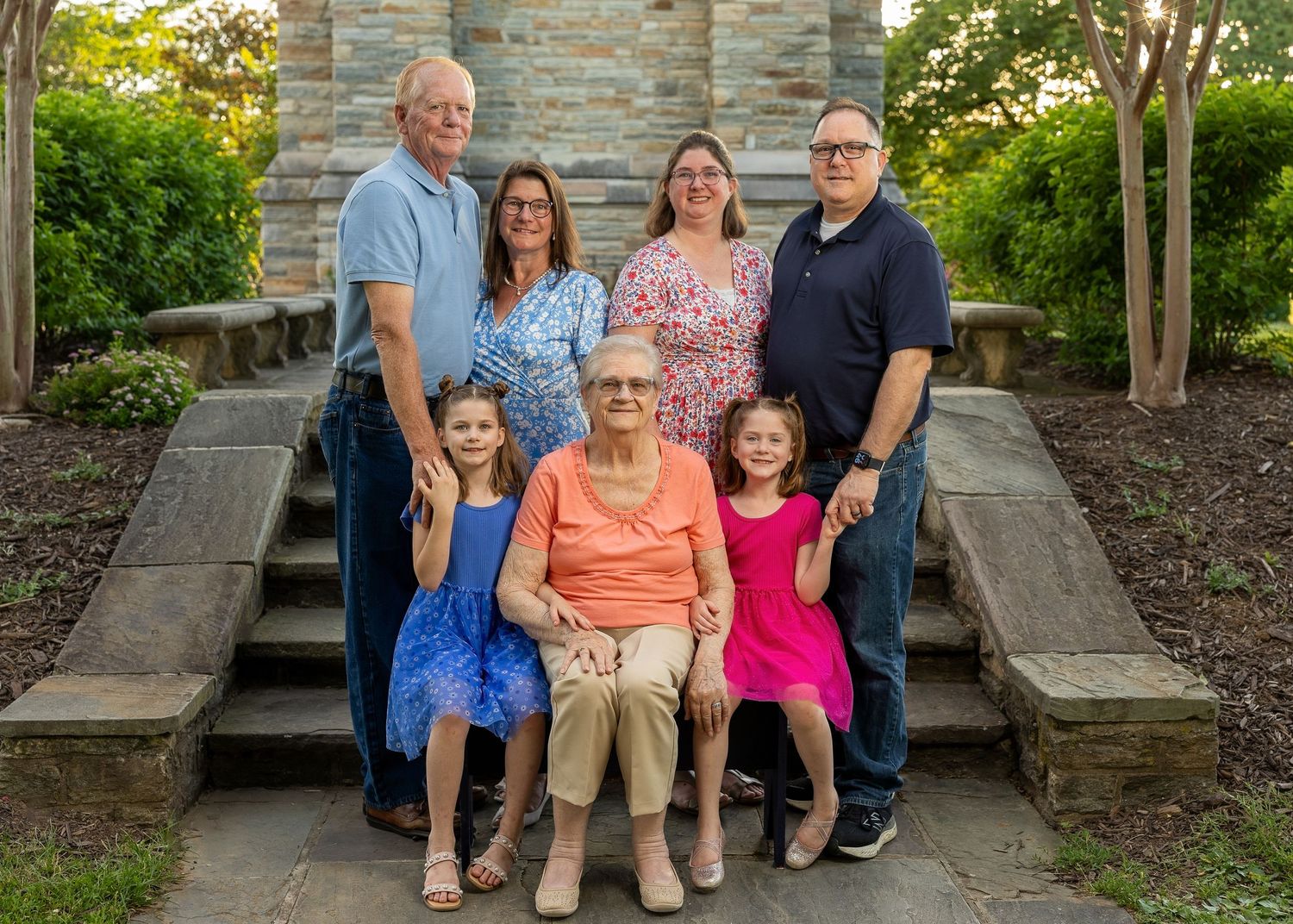 Multi-generational family portrait with seven members posing on stone steps surrounded by lush greenery outdoors.