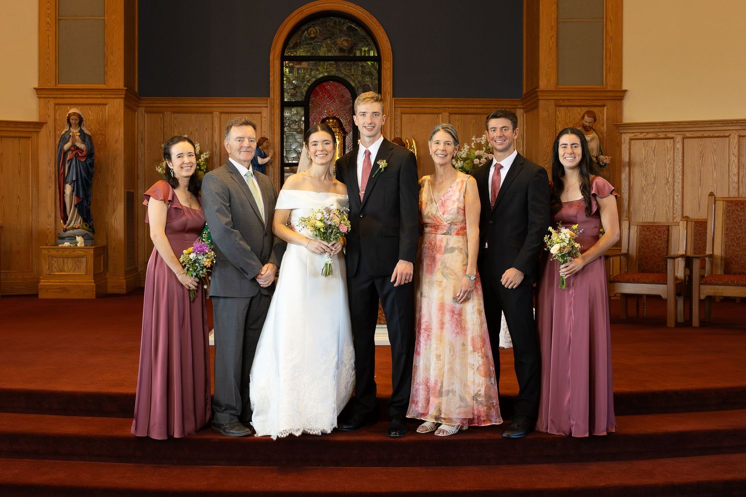 Wedding party poses in a church, bride in white gown with groom and bridesmaids in mauve dresses before altar.