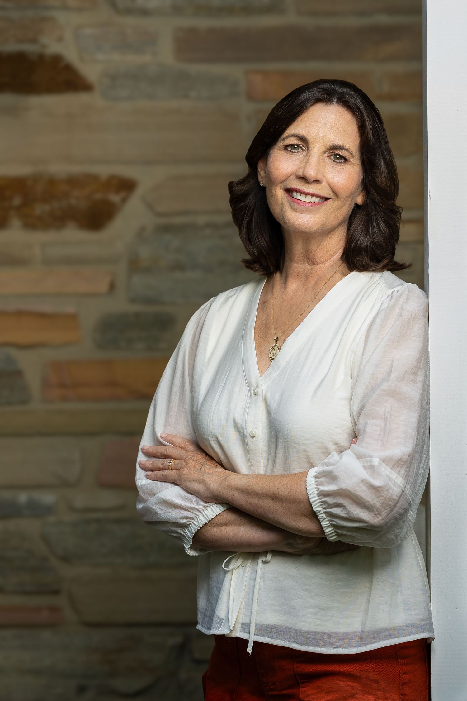 Smiling middle-aged woman with dark hair in white blouse and red pants, arms crossed, leaning against stone wall.