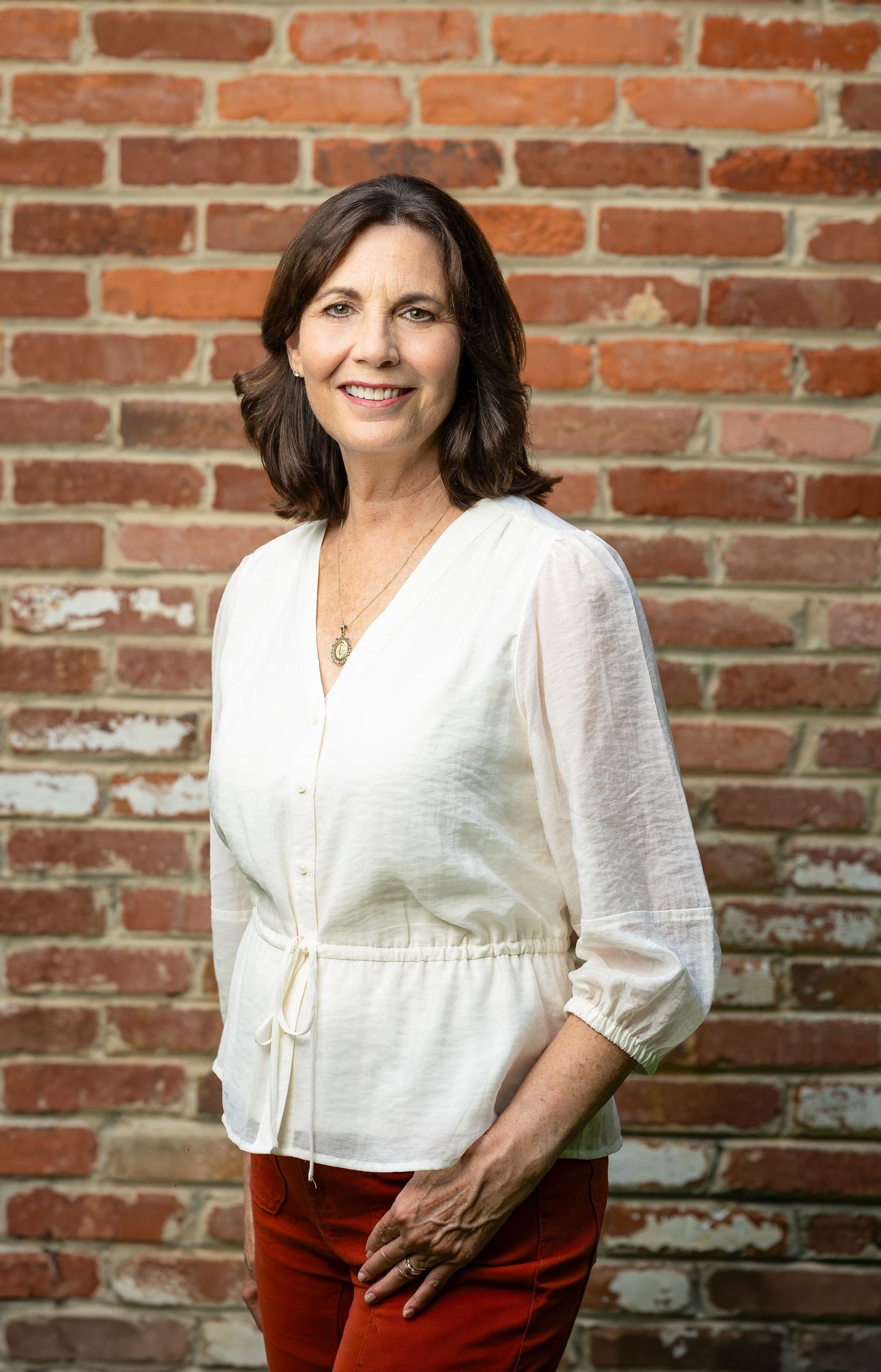 Smiling woman in white blouse and red pants posing confidently against a rustic brick wall backdrop.