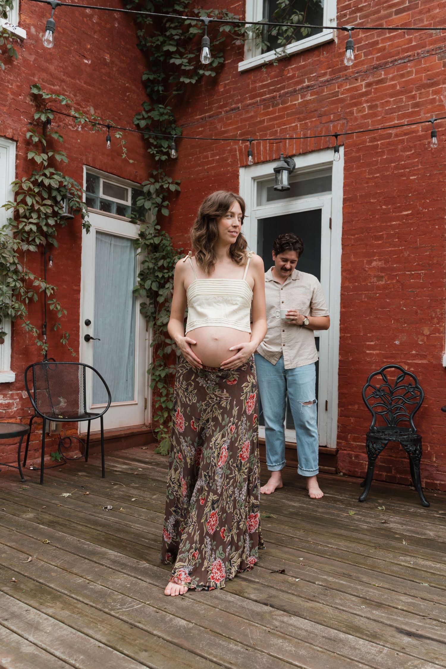 Pregnant woman in floral skirt holds baby bump on wooden deck outside red brick home with man in background.