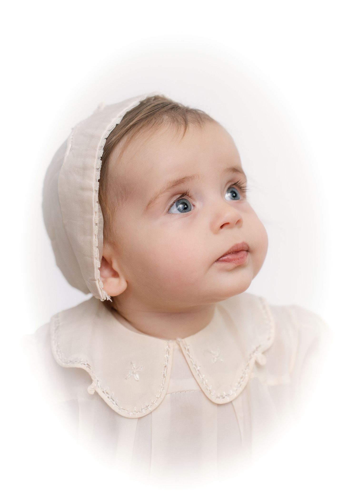 Adorable baby girl with blue eyes wearing a cream bonnet and scalloped collar dress.
