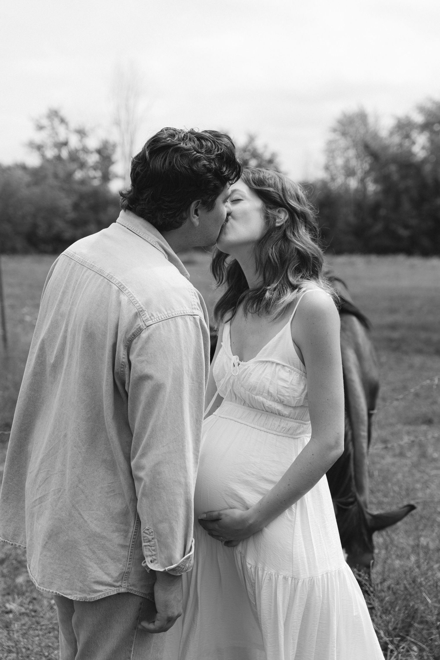 Black and white photo of a pregnant couple kissing in a field with a horse in the background.