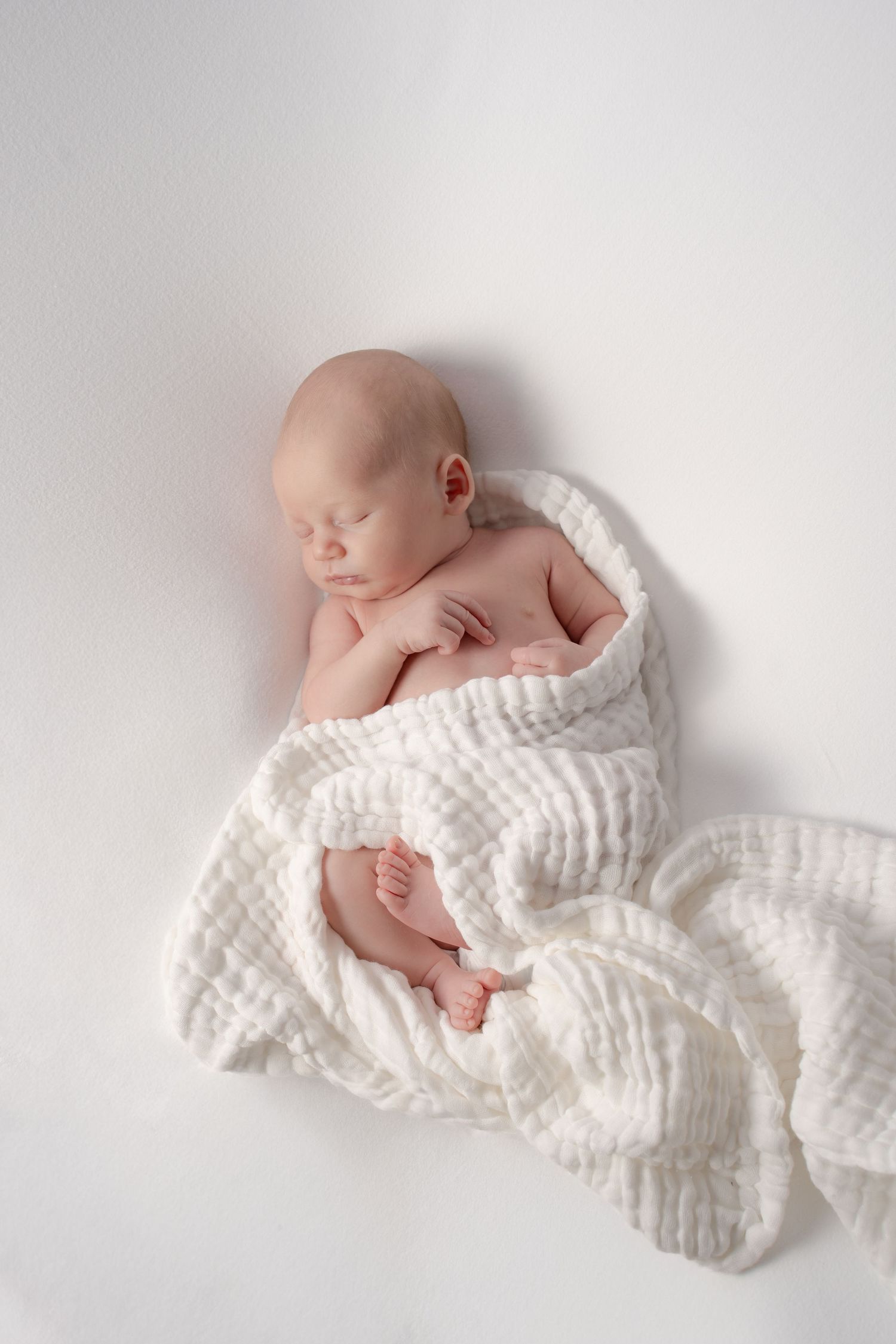Peaceful newborn baby sleeping wrapped in a soft white textured blanket on a white background.