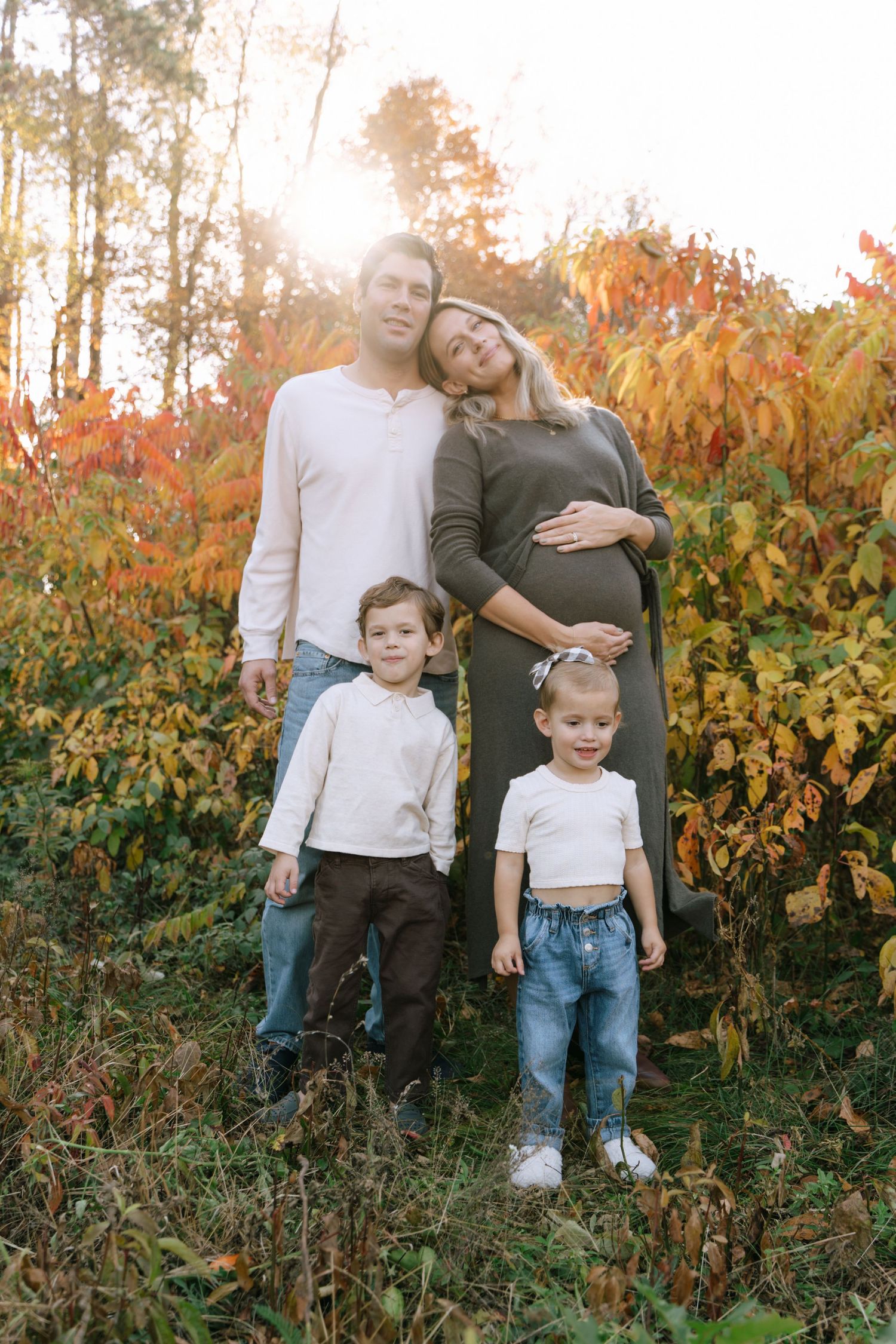 Pregnant woman and husband pose with two young children in autumn forest with golden sunlight filtering through colorful fall foliage.
