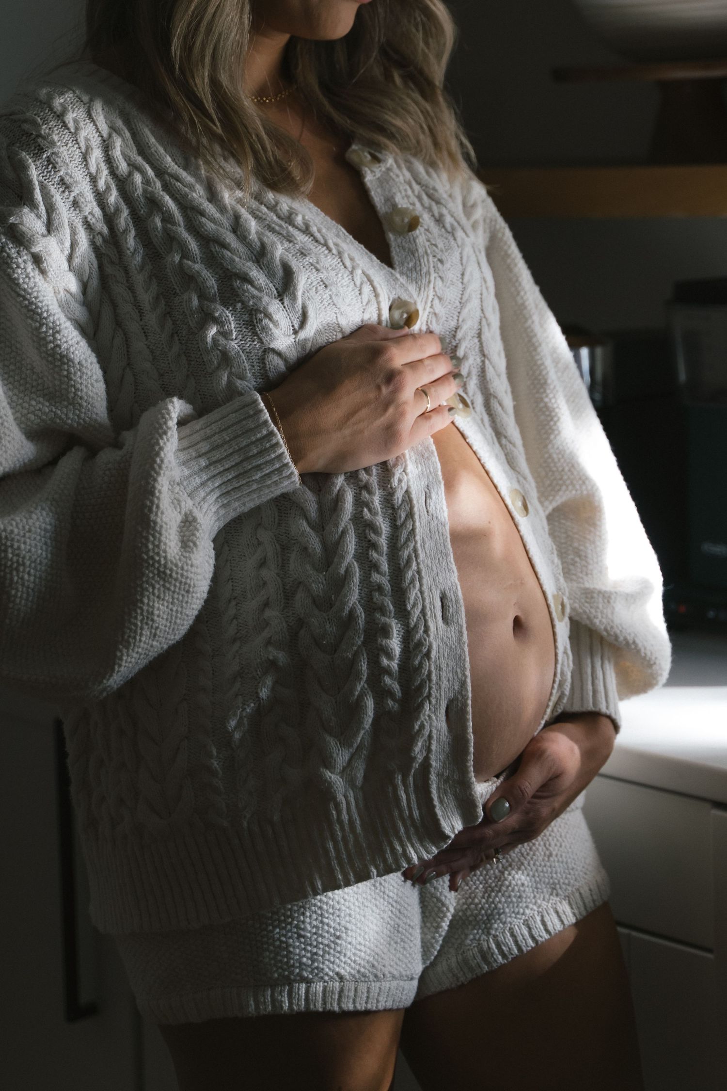Pregnant woman in white cable-knit cardigan and shorts cradling baby bump, soft natural light.