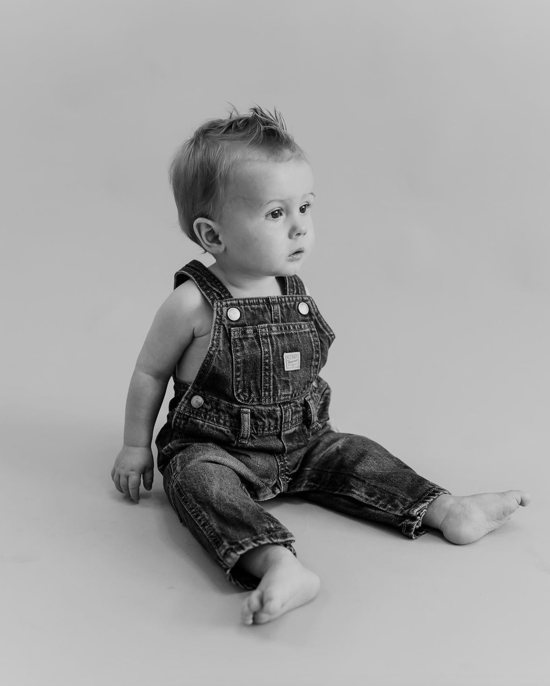 Black and white photo of a baby boy sitting on the floor wearing denim overalls.