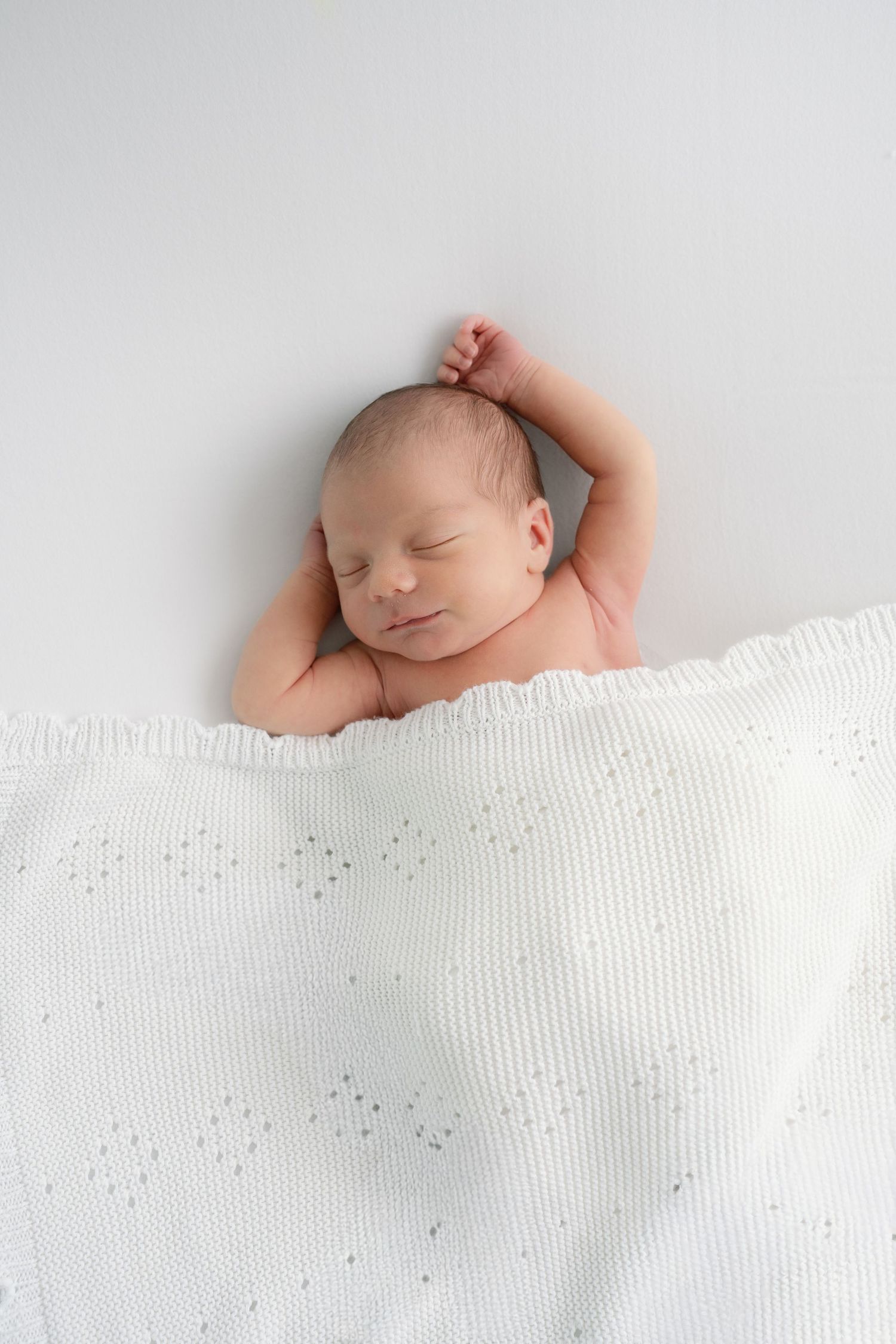 Peaceful sleeping newborn baby covered with white knit blanket on light gray background.