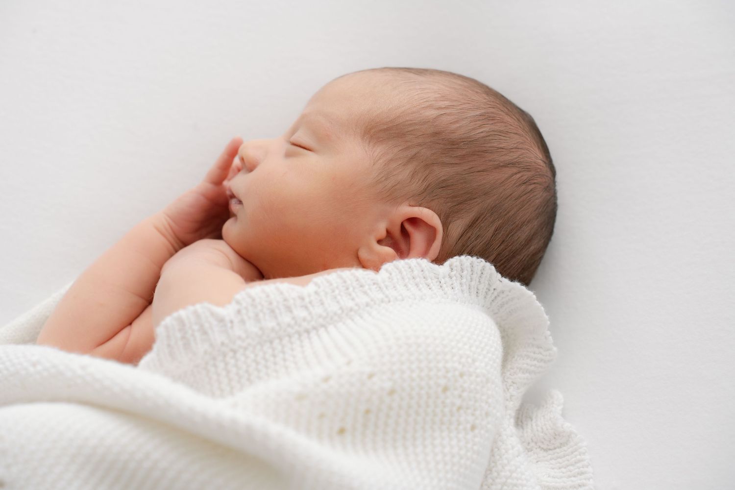Close-up of sleeping newborn baby wrapped in soft white knit blanket on white surface.