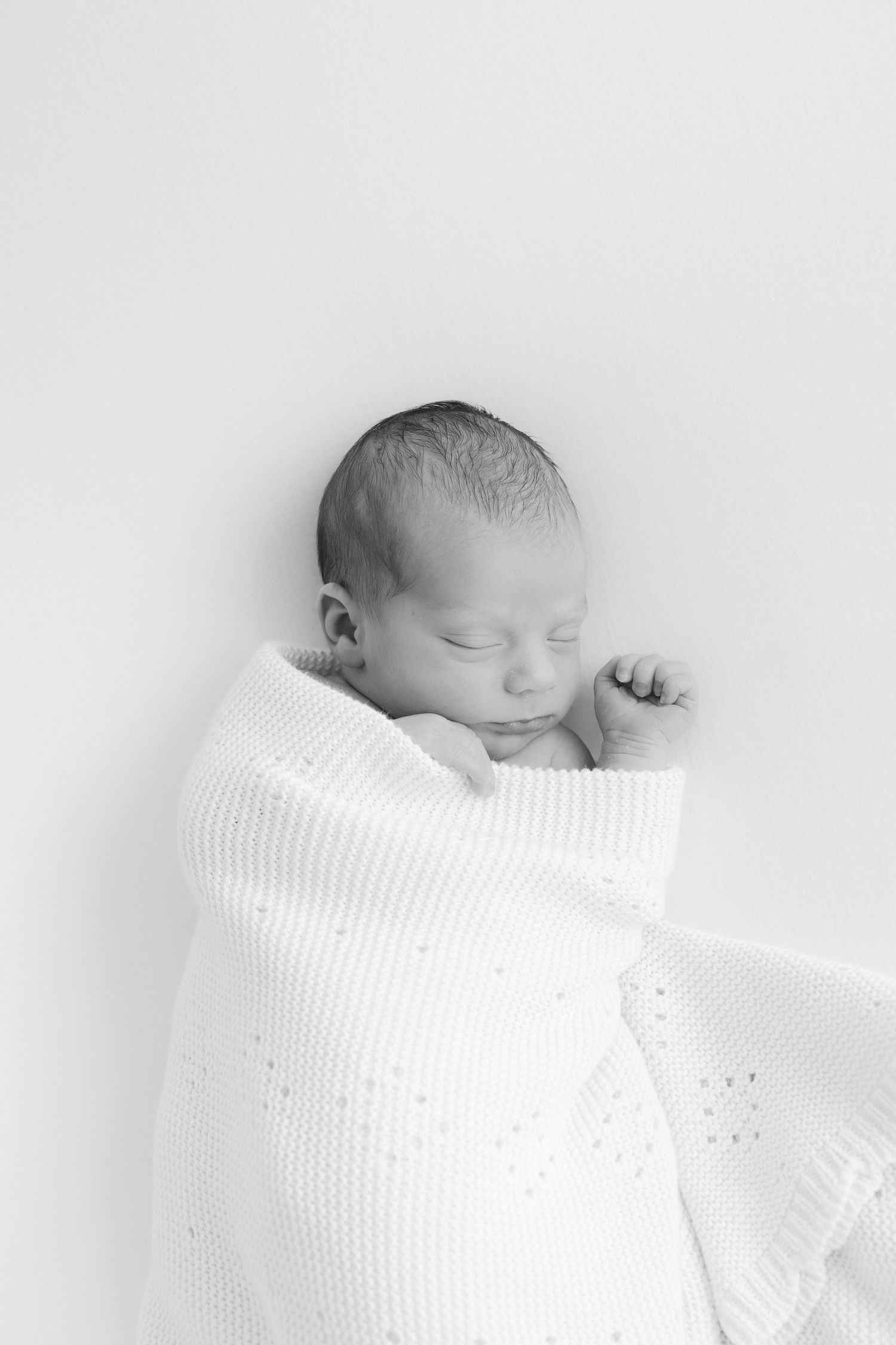 Sleeping newborn baby swaddled in a white knit blanket, black and white portrait photography.