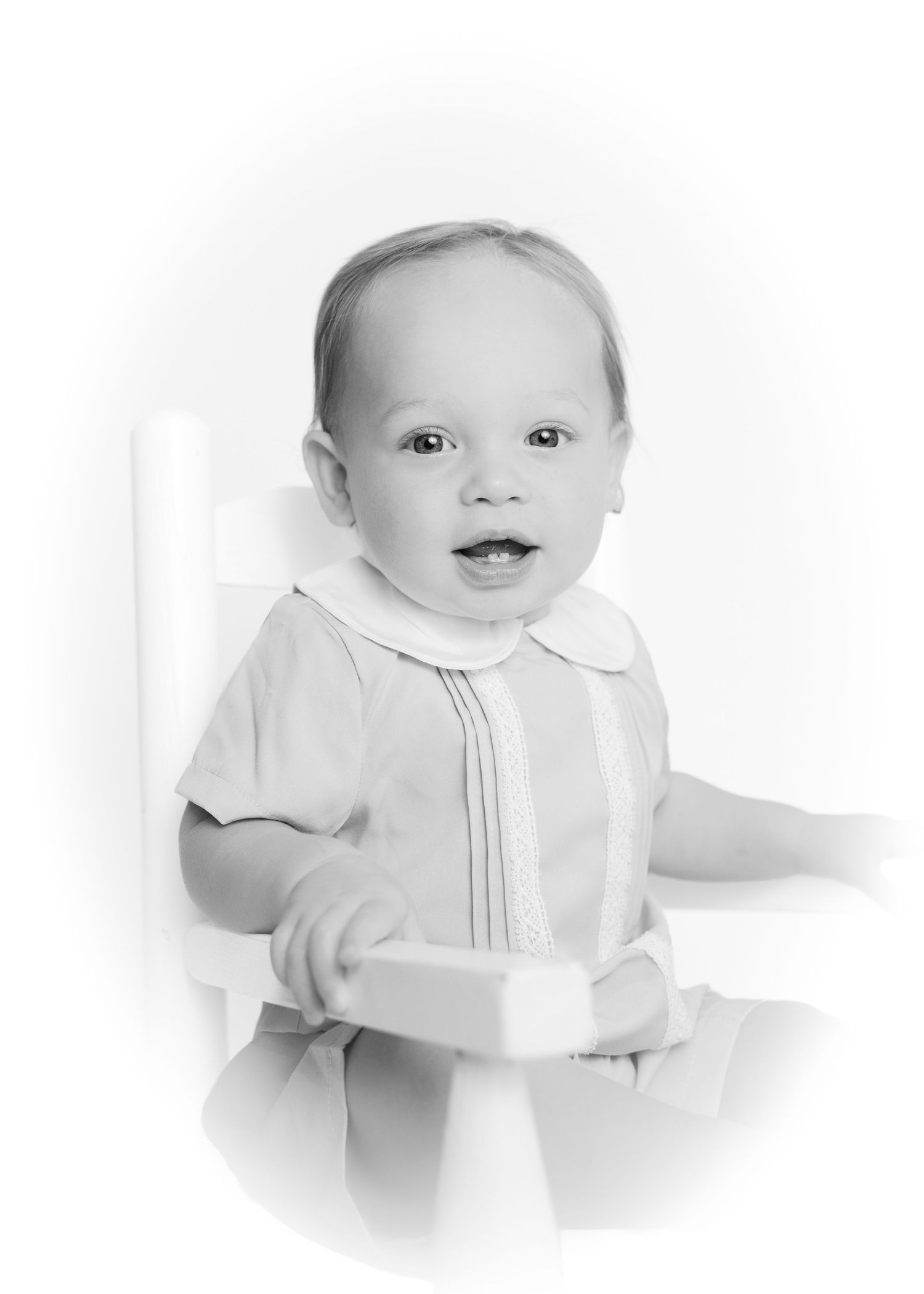 Black and white portrait of smiling baby boy sitting in white chair with vignette.