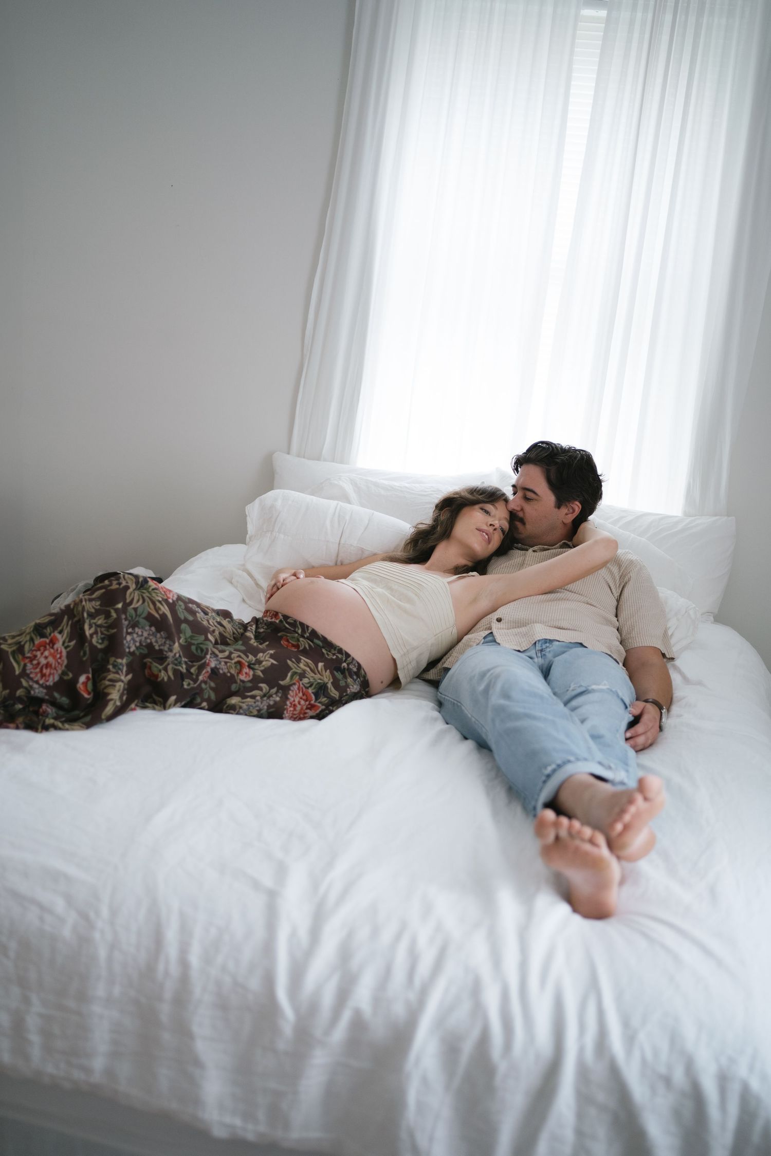 Pregnant couple relaxing together on a white bed, man kissing woman's head in a bright bedroom.