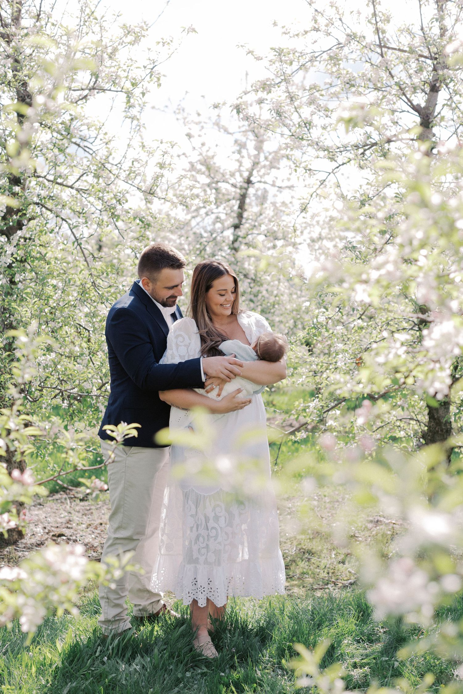 Happy couple holding newborn baby in blooming spring orchard with white flowers.