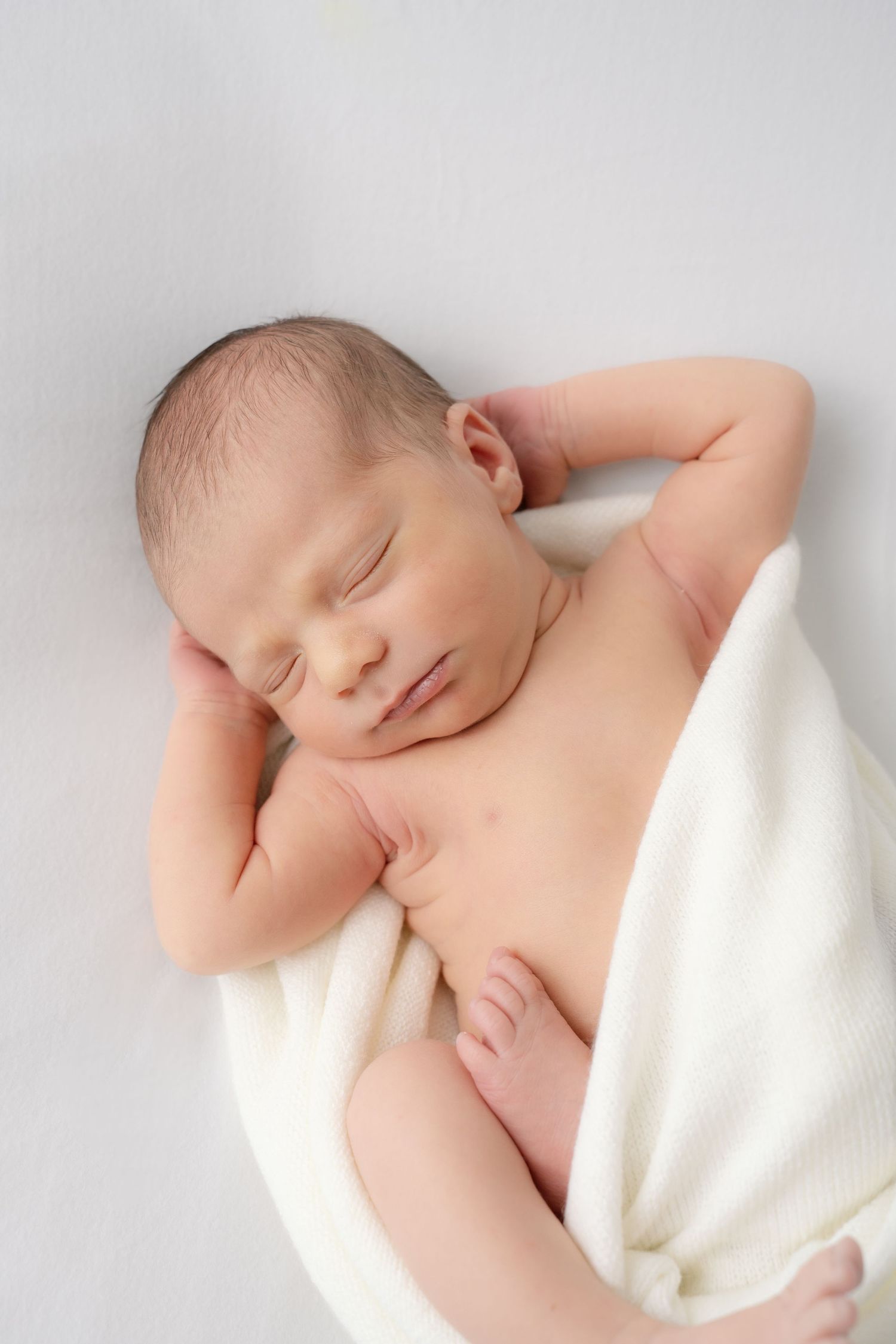 Peaceful newborn baby sleeping wrapped in a soft white blanket with tiny hands near face.