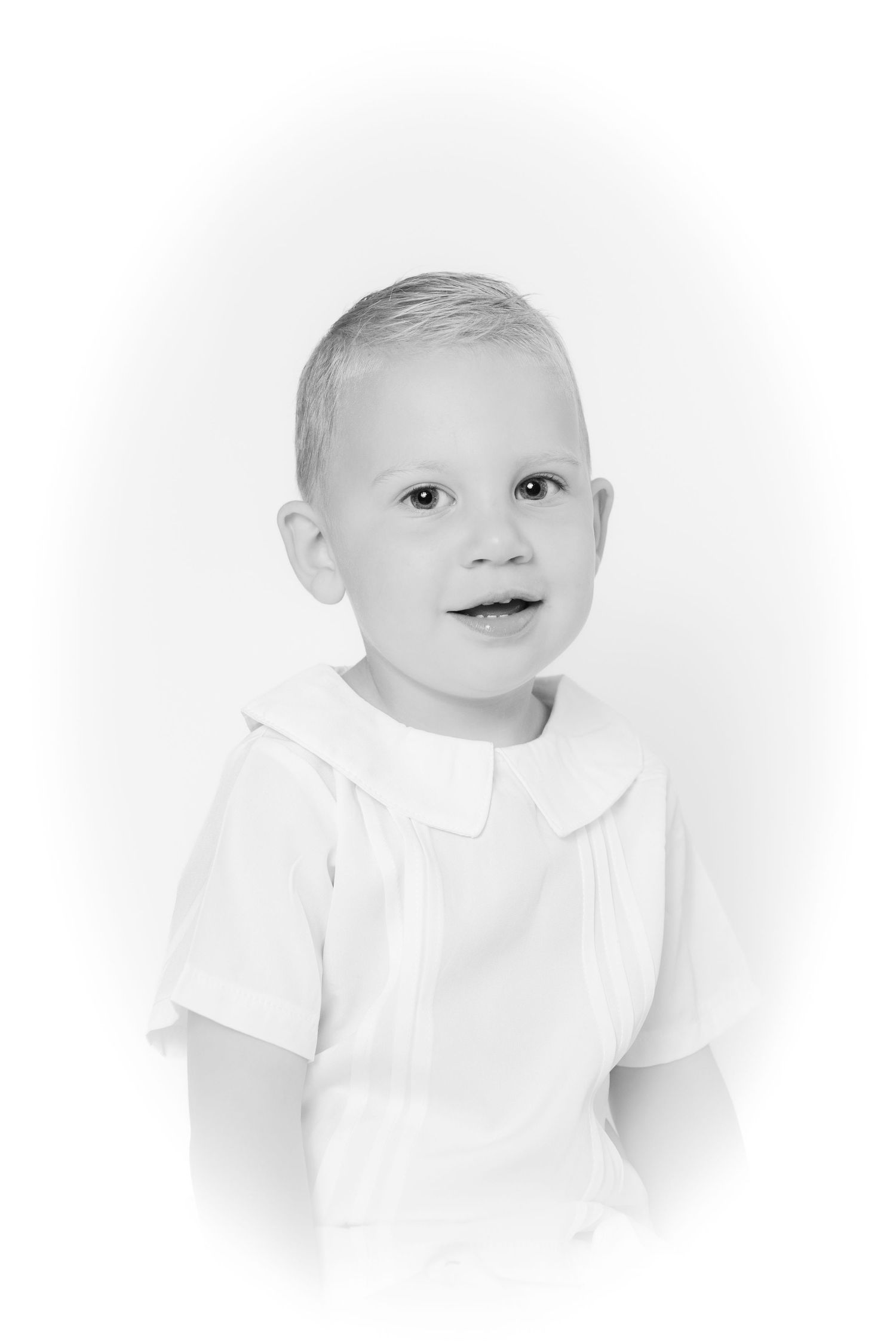 Black and white vignette portrait of young blonde boy in white collared shirt smiling.