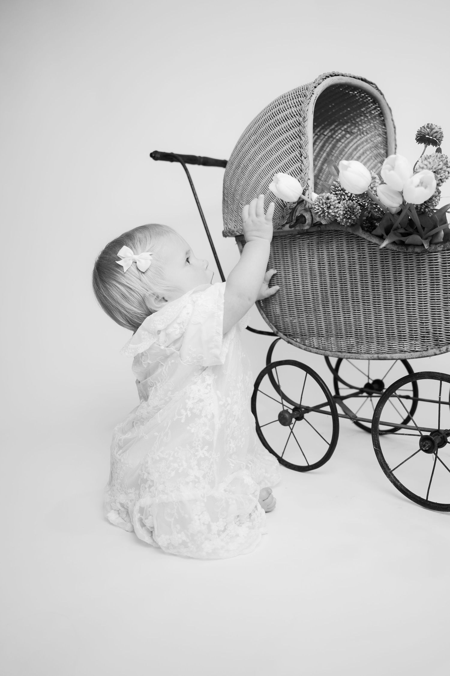 Black and white photo of baby girl in lace dress reaching up to vintage wicker pram.