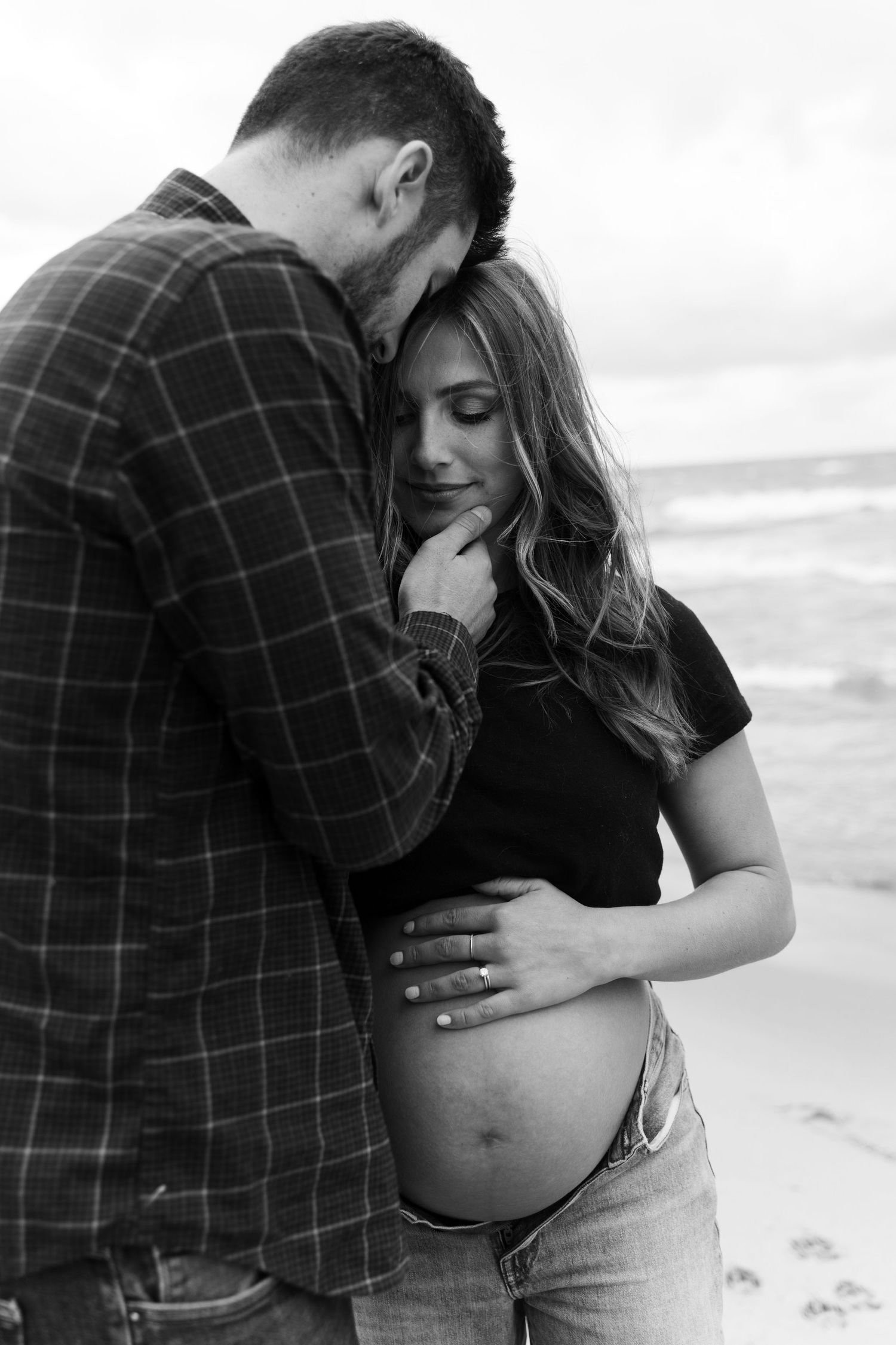 Black and white maternity photo of a couple embracing on a beach, man kissing woman's head while she cradles her baby bump.