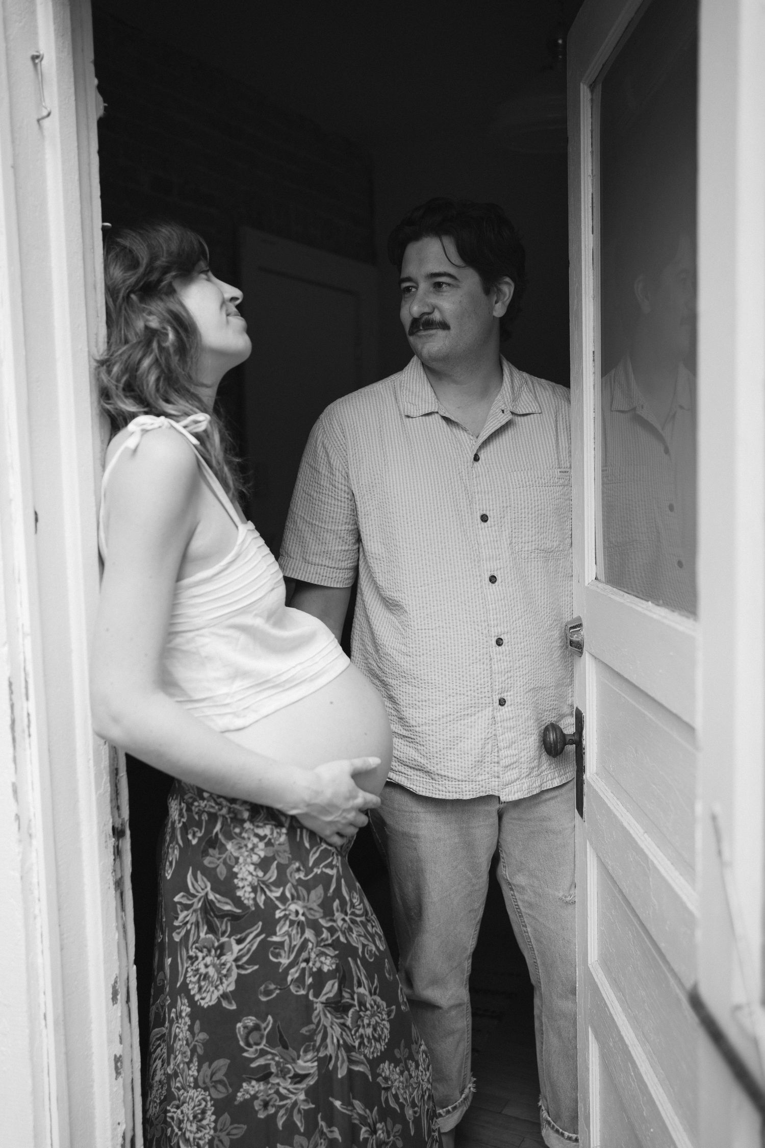 Black and white photo of pregnant couple sharing a smile in a rustic doorway.