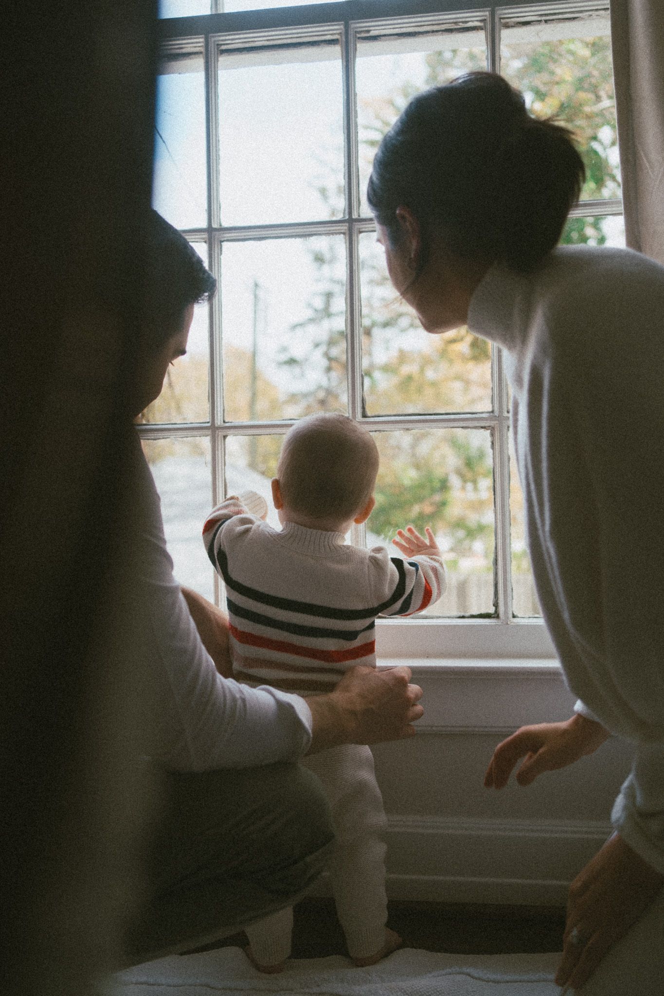 Parents with baby in striped outfit looking out a sunlit window together.