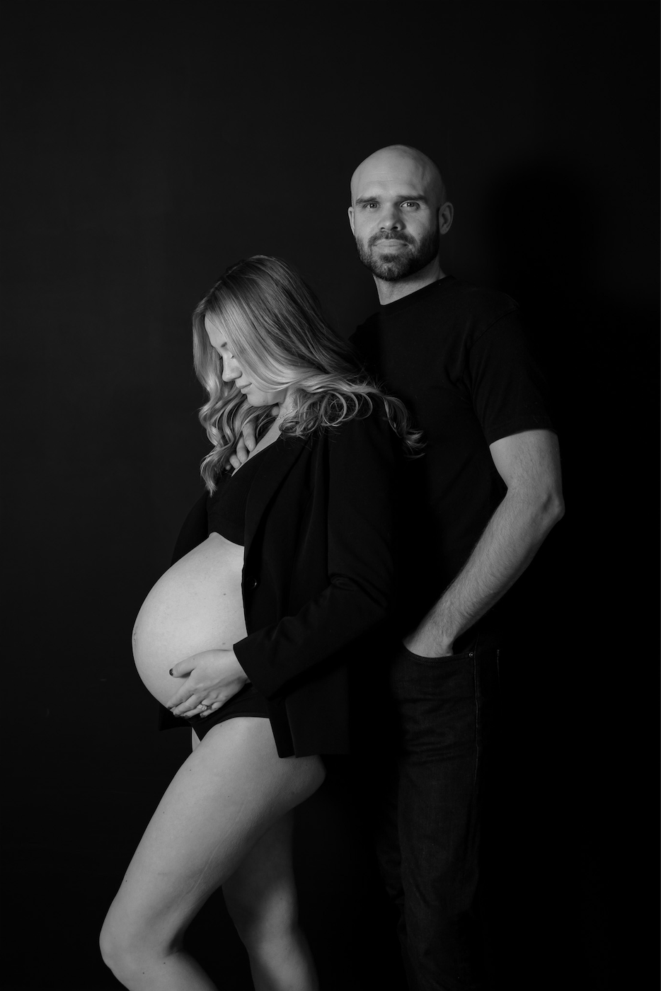 Black and white maternity photo of a pregnant woman and man posing together against a dark background.