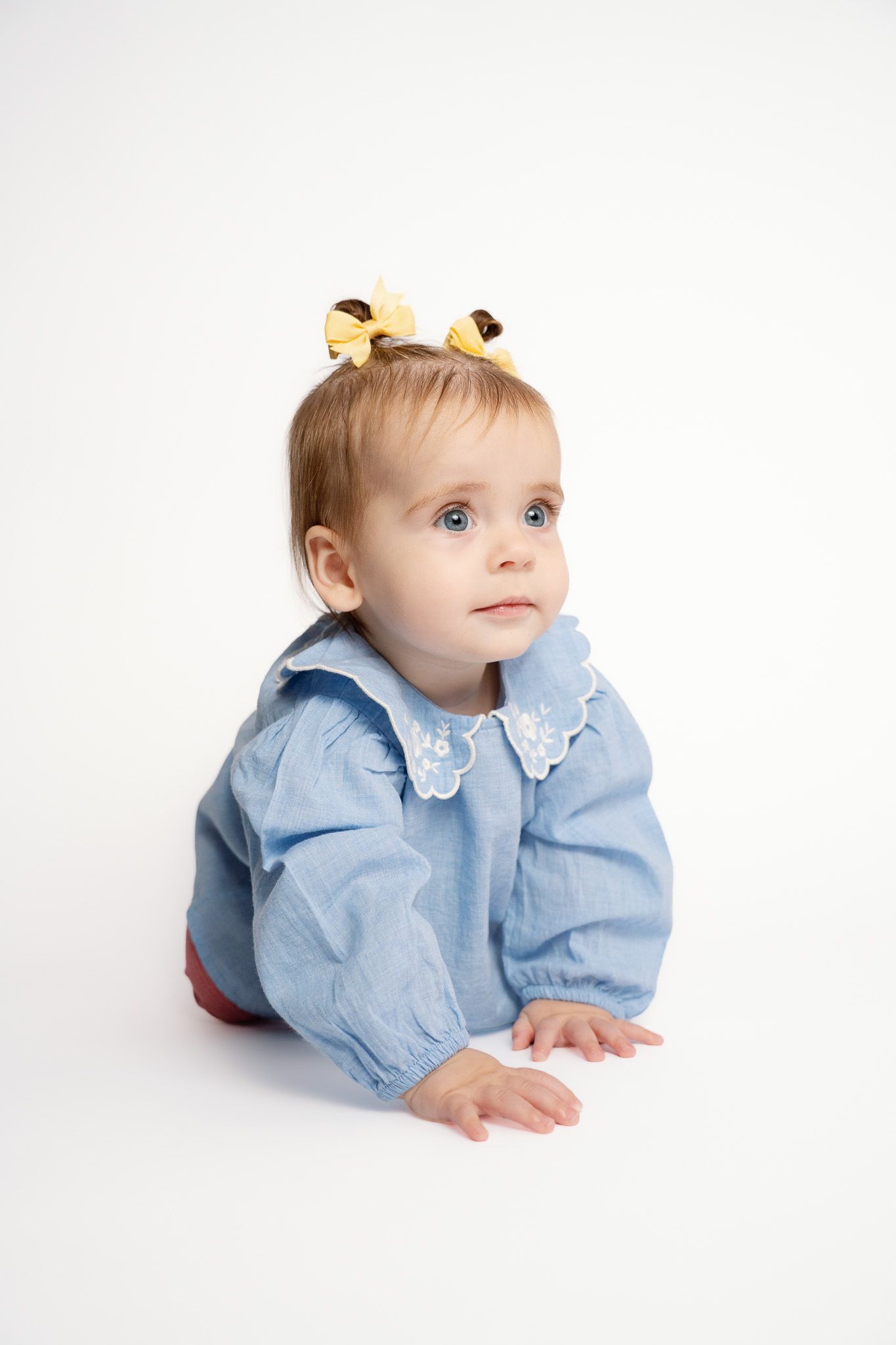 Baby girl in blue blouse with white collar and yellow hair bows, looking up curiously on white background photographed in Rockford Michigan