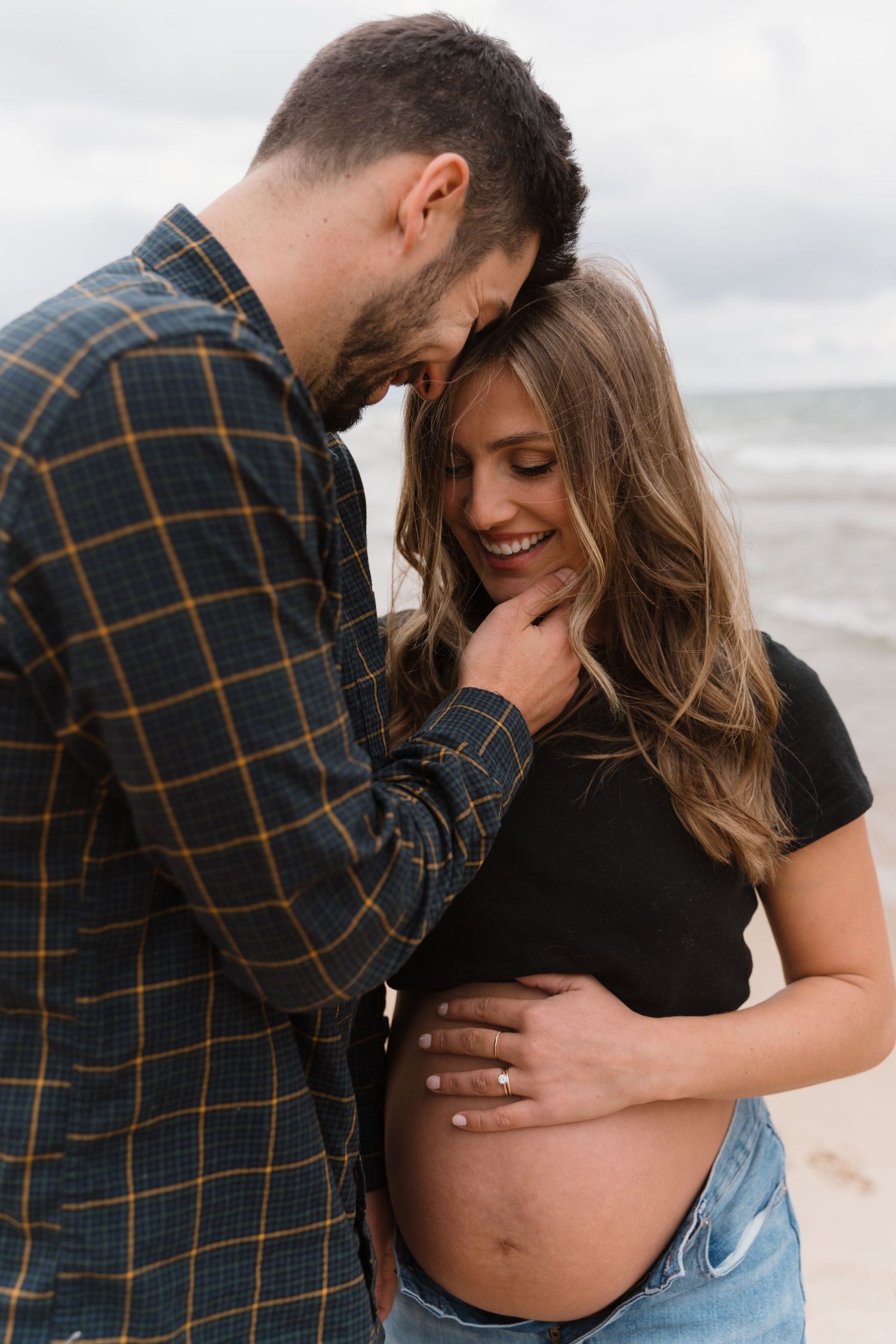 Couple at beach, man tenderly touching pregnant womans chin as she smiles at bump.