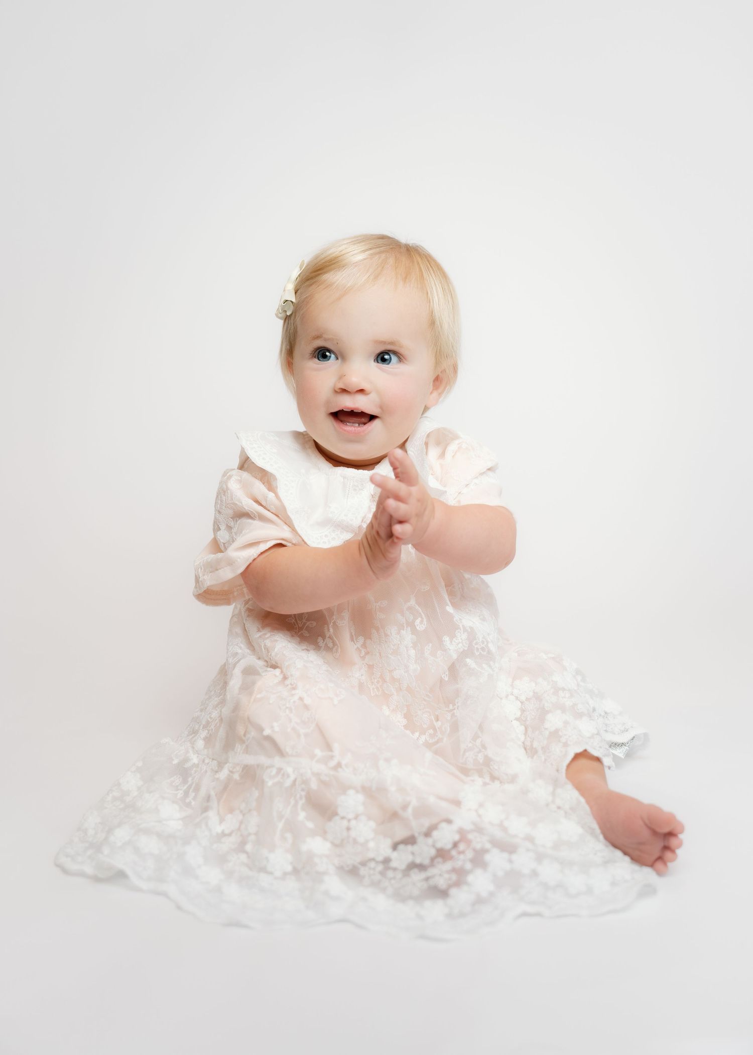 Smiling baby girl in white lace dress sitting up, with blonde hair and blue eyes, on white background.