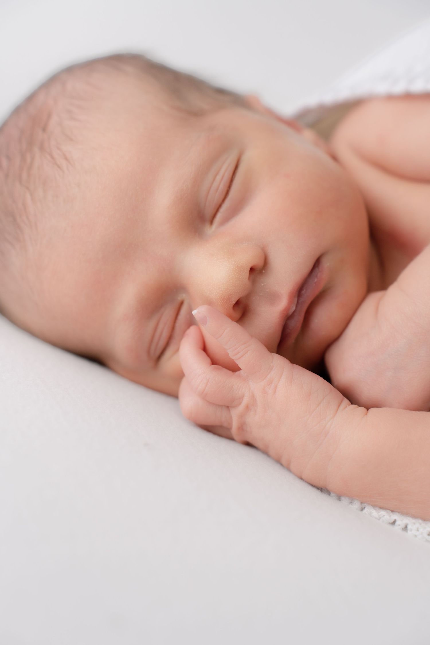 Close-up of a sleeping newborn baby's face with tiny fingers touching their lips on white.