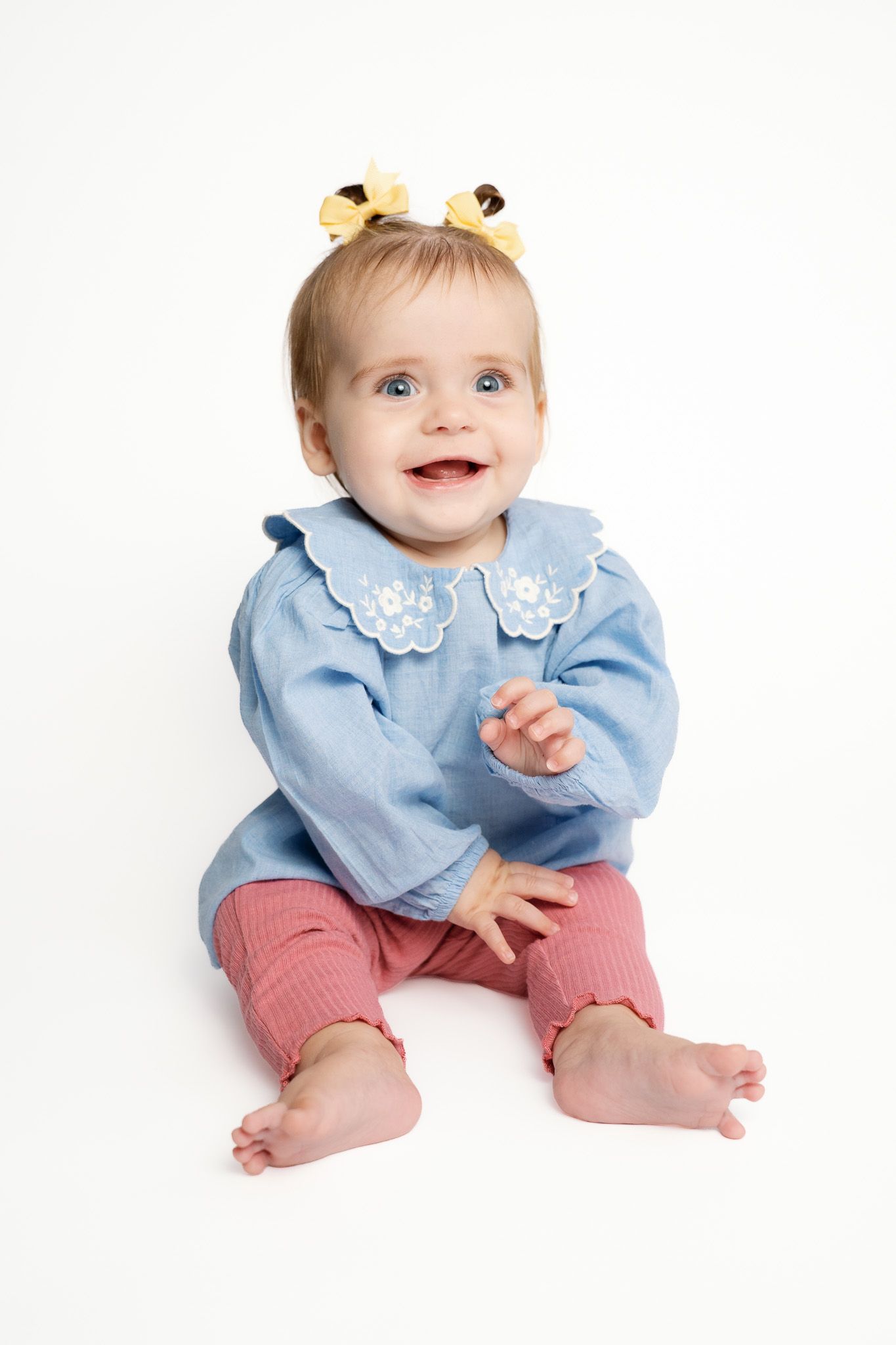 Smiling baby girl in blue top and pink pants with yellow hair bows sits barefoot on white background for a milestone portrait in Michigan