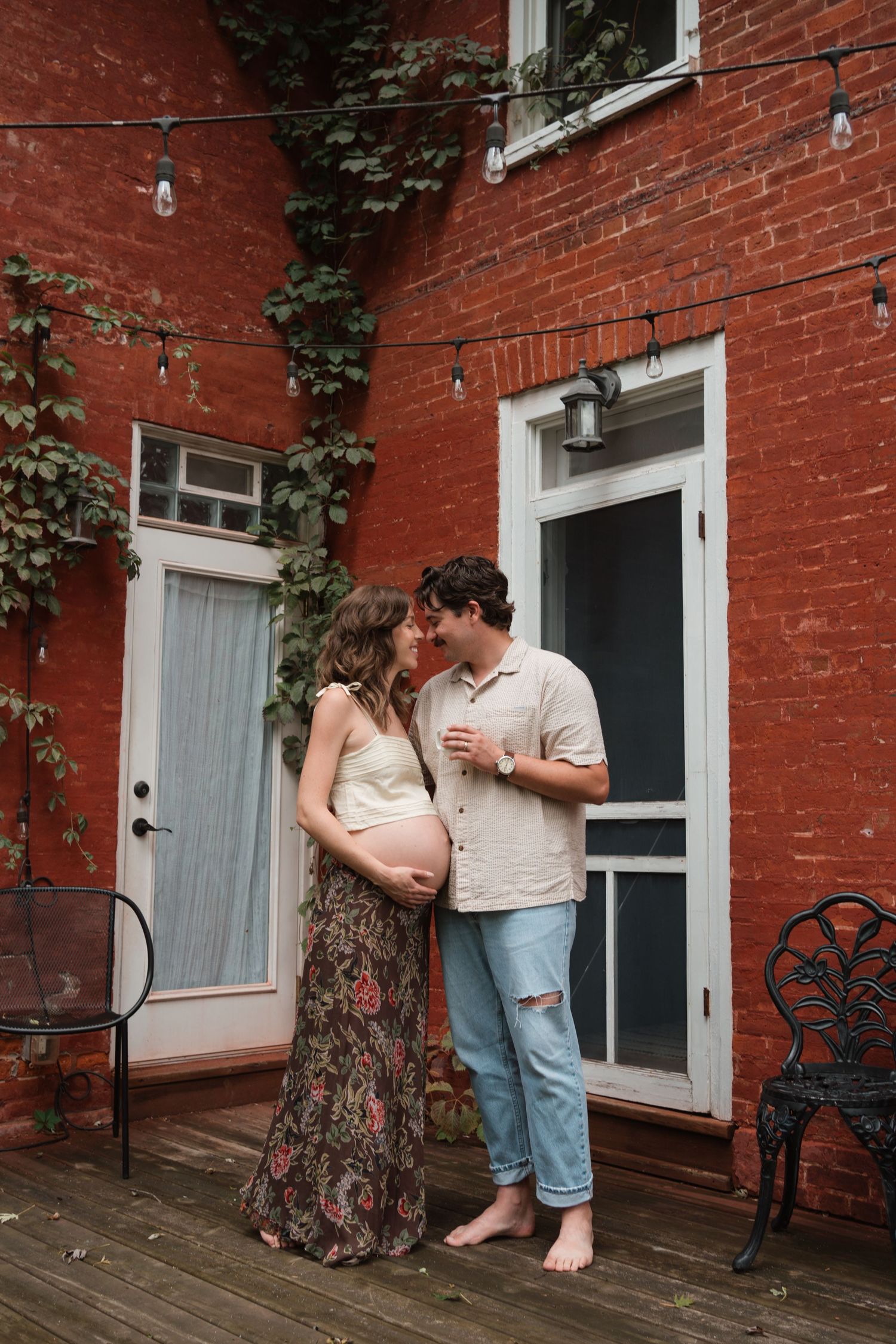 Couple sharing a tender kiss on a wooden deck outside a red brick building, woman in floral skirt cradling her pregnant belly.