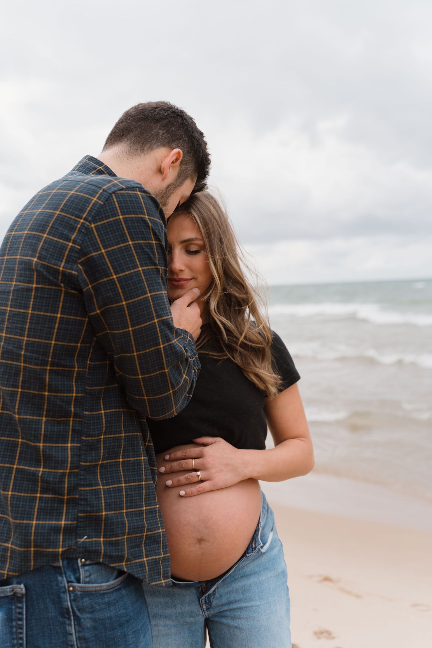 Couple embracing on a cloudy beach, woman showing her pregnant belly outdoors.