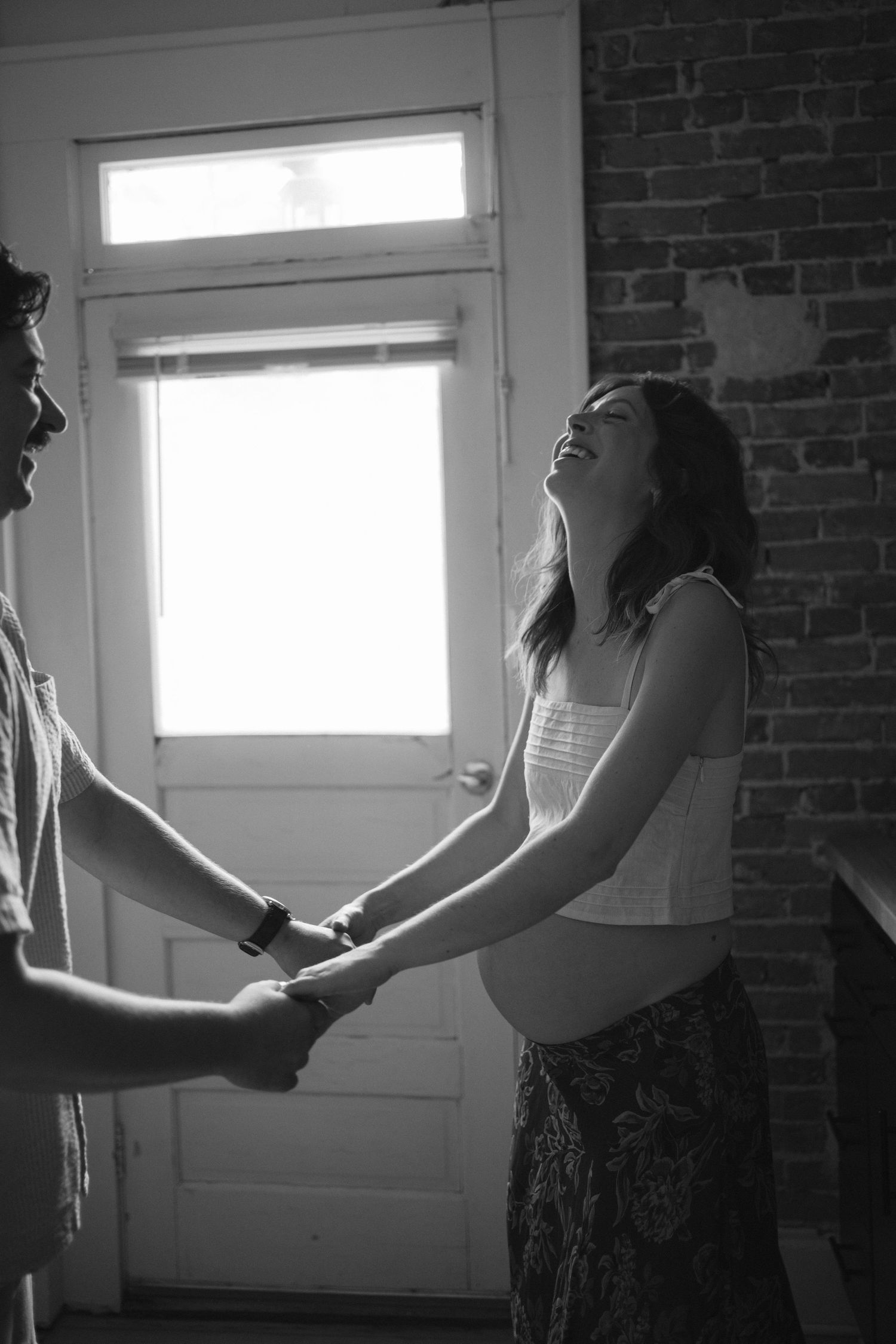 Black and white photo of laughing pregnant woman holding hands with partner near bright window.