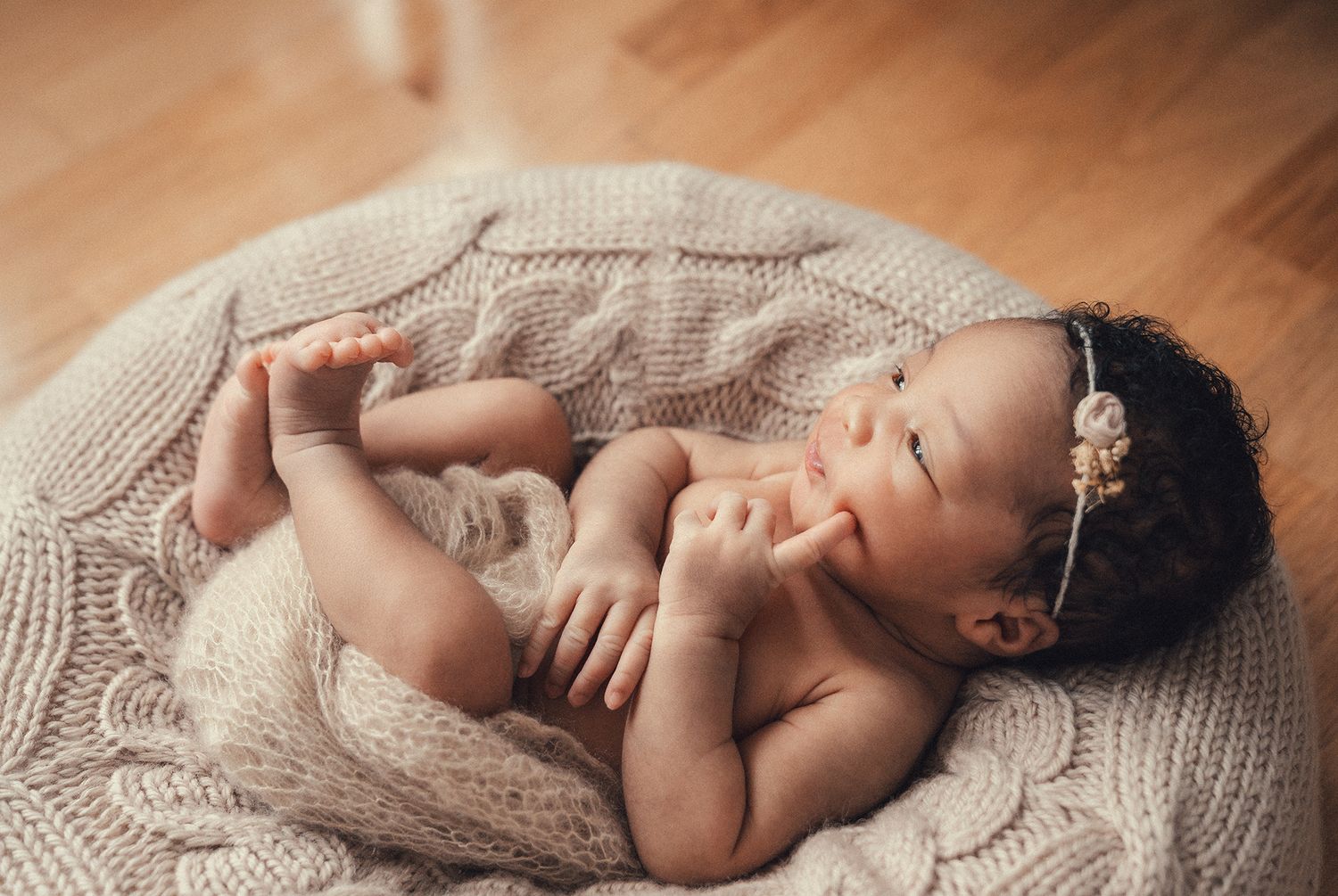 Adorable newborn baby girl with a floral headband resting in a knit bowl wrap on a wooden floor.