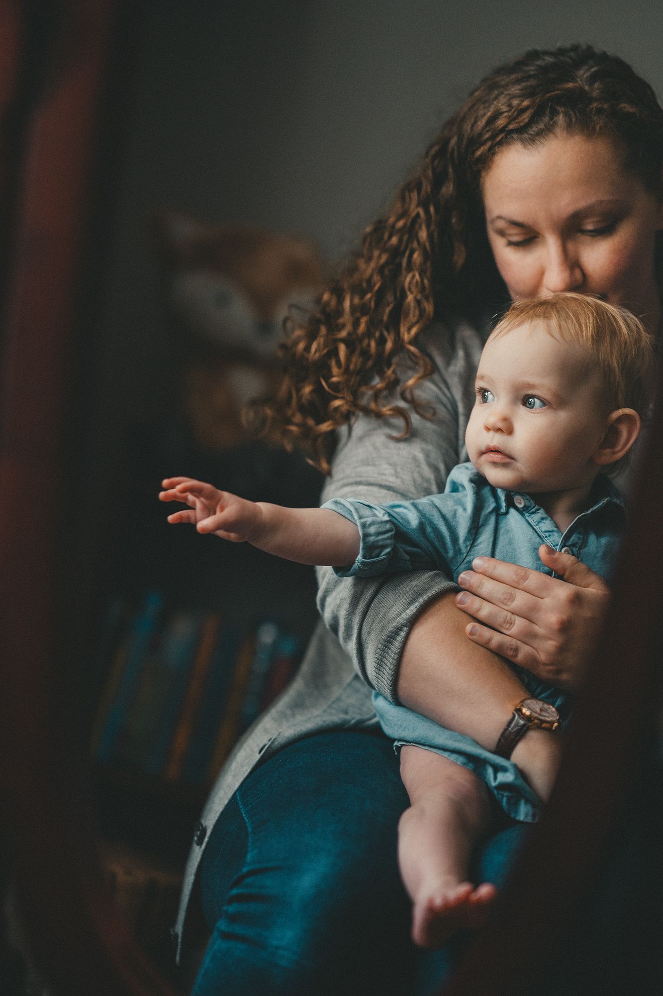 A curly-haired mother tenderly holds her blue-eyed baby boy in a denim shirt indoors.