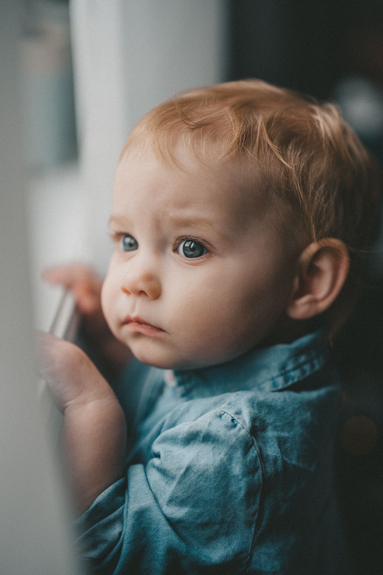 A curious red-haired baby gazes out a window, gently touching the glass with tiny fingers.