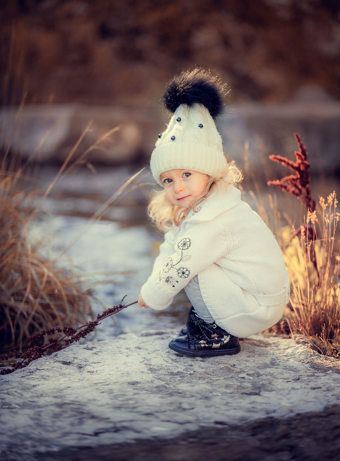 A smiling toddler girl in a white winter coat and pom-pom hat crouches on a snowy surface outdoors.