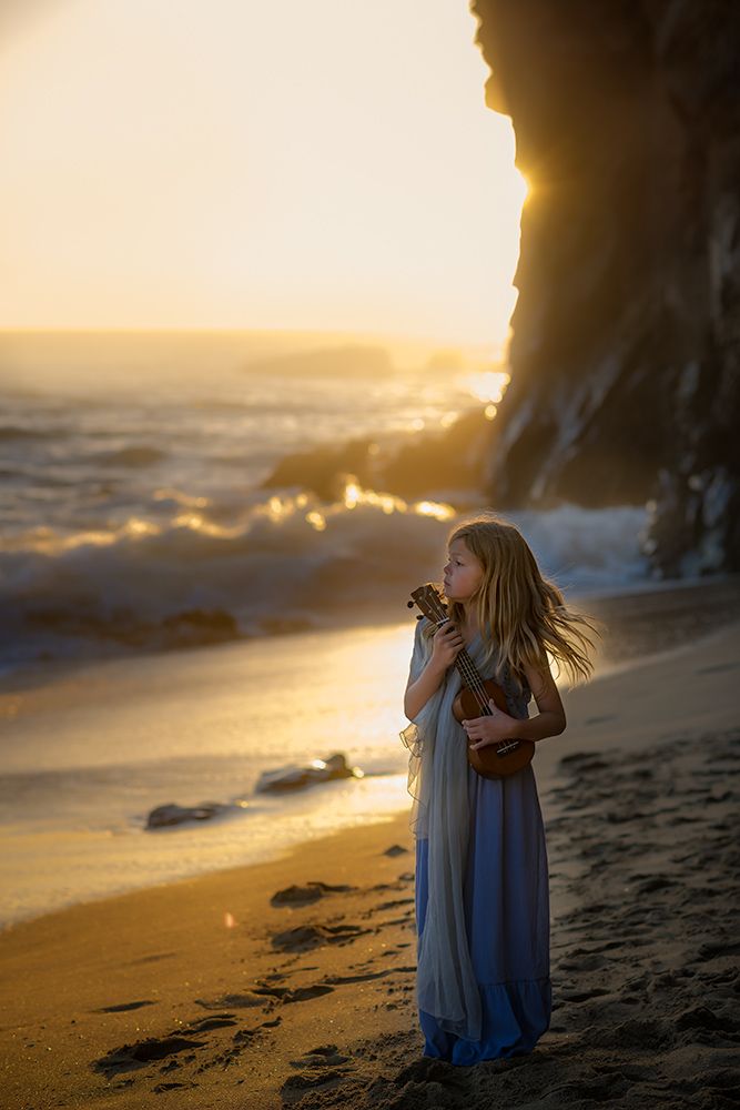 A young girl in a flowing blue dress holds a ukulele while standing on a beach at golden sunset near rocky cliffs.