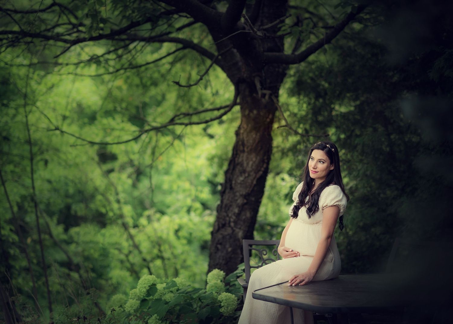 Pregnant woman in white dress sitting on bench surrounded by lush green trees.