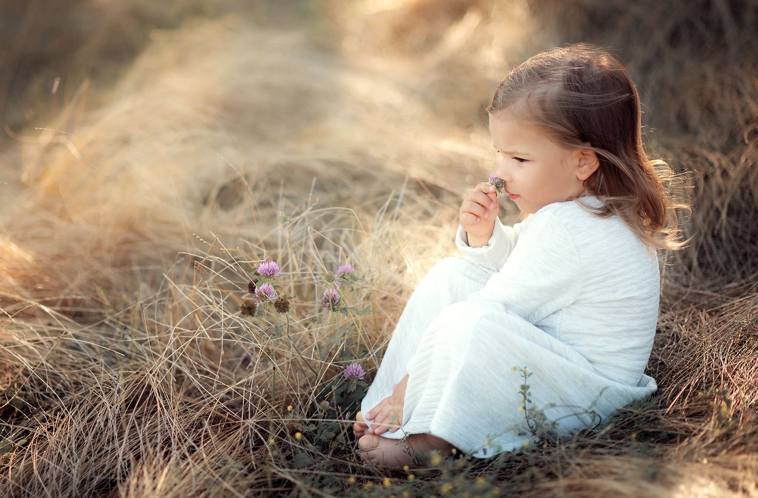 Little girl in white dress sitting peacefully in dry golden grass field outdoors.
