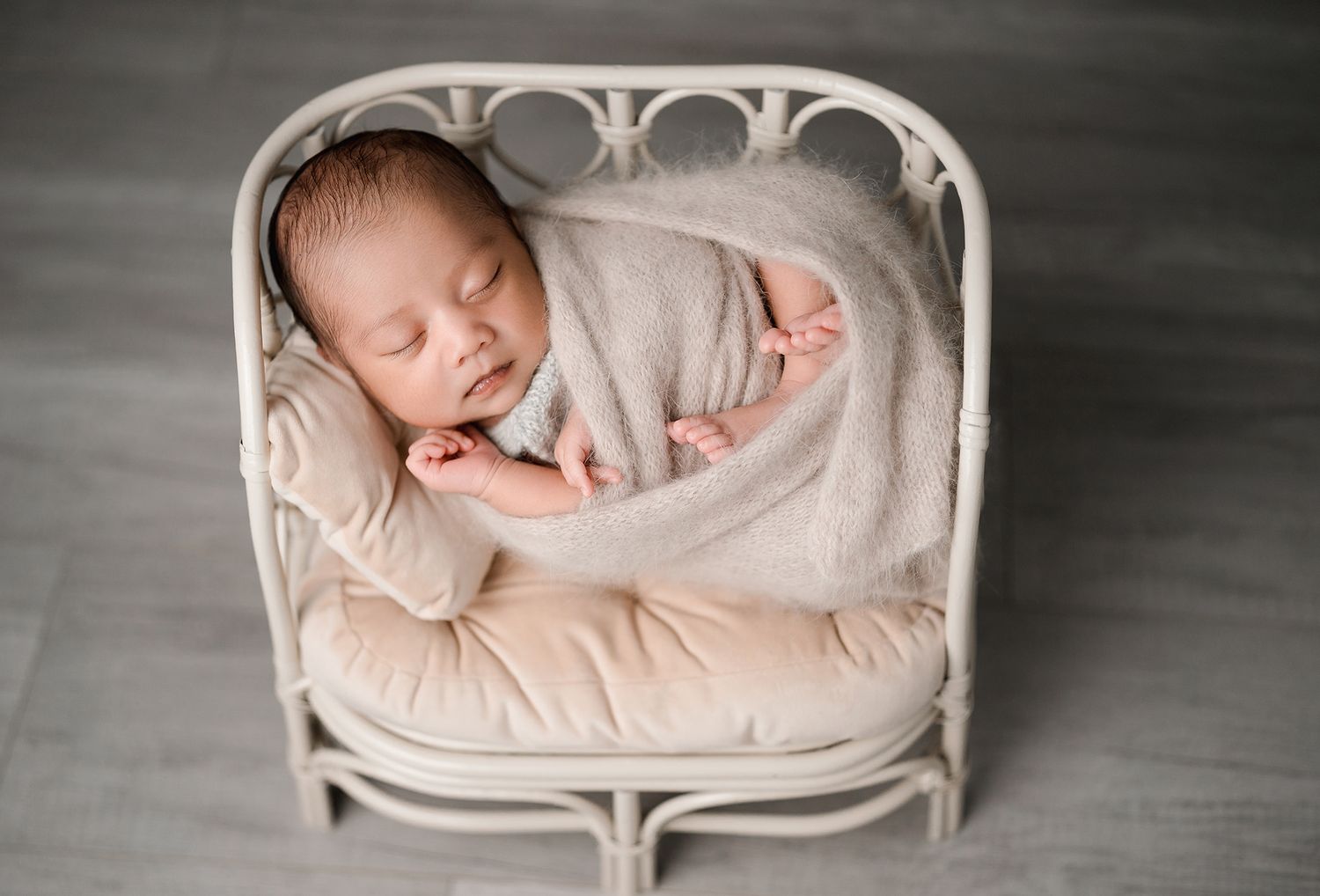 Newborn baby sleeping peacefully, wrapped in a soft beige blanket and resting in a small white rattan chair with a cushion.