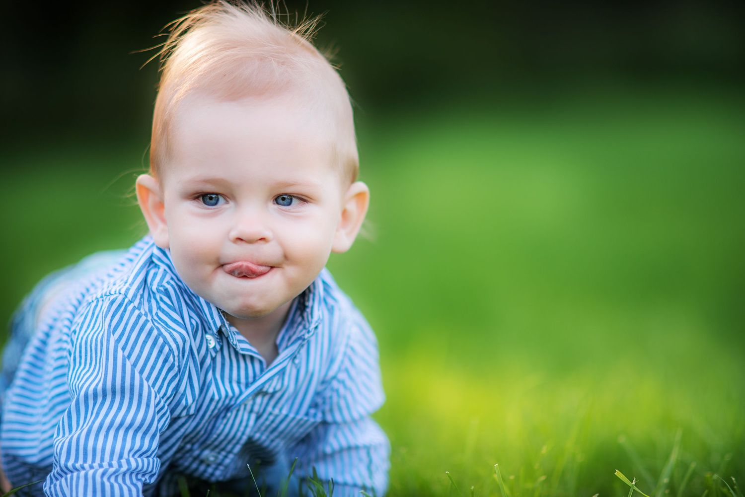 Playful baby boy in striped shirt crawling on green grass sticking tongue out.