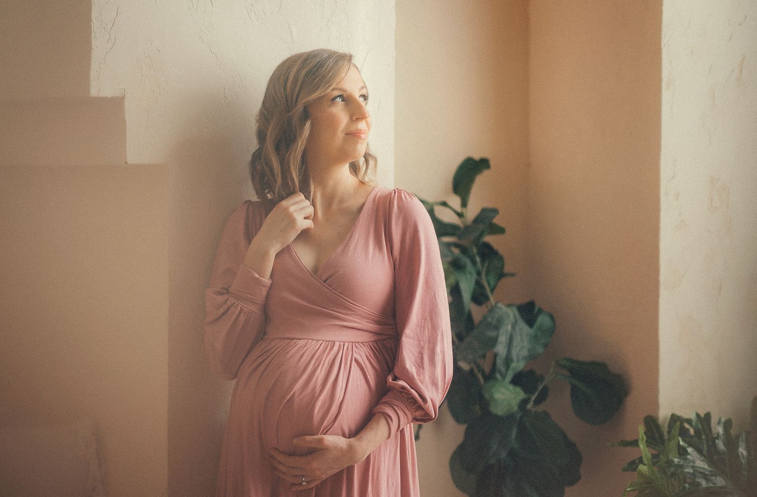 Pregnant woman in pink maxi dress cradling baby bump by sunlit window with green plant.