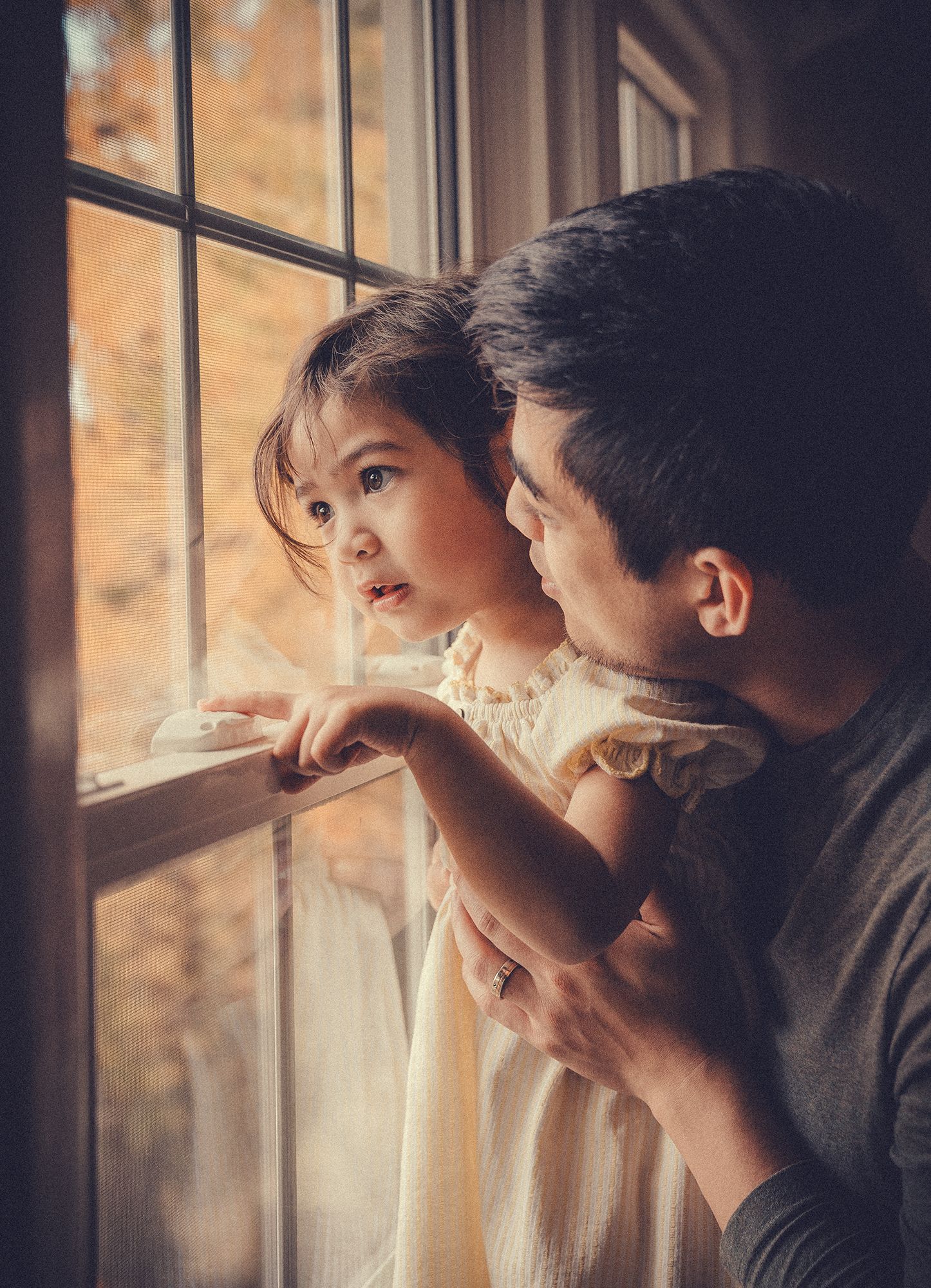 Father and toddler daughter looking out a window together in warm autumn light.