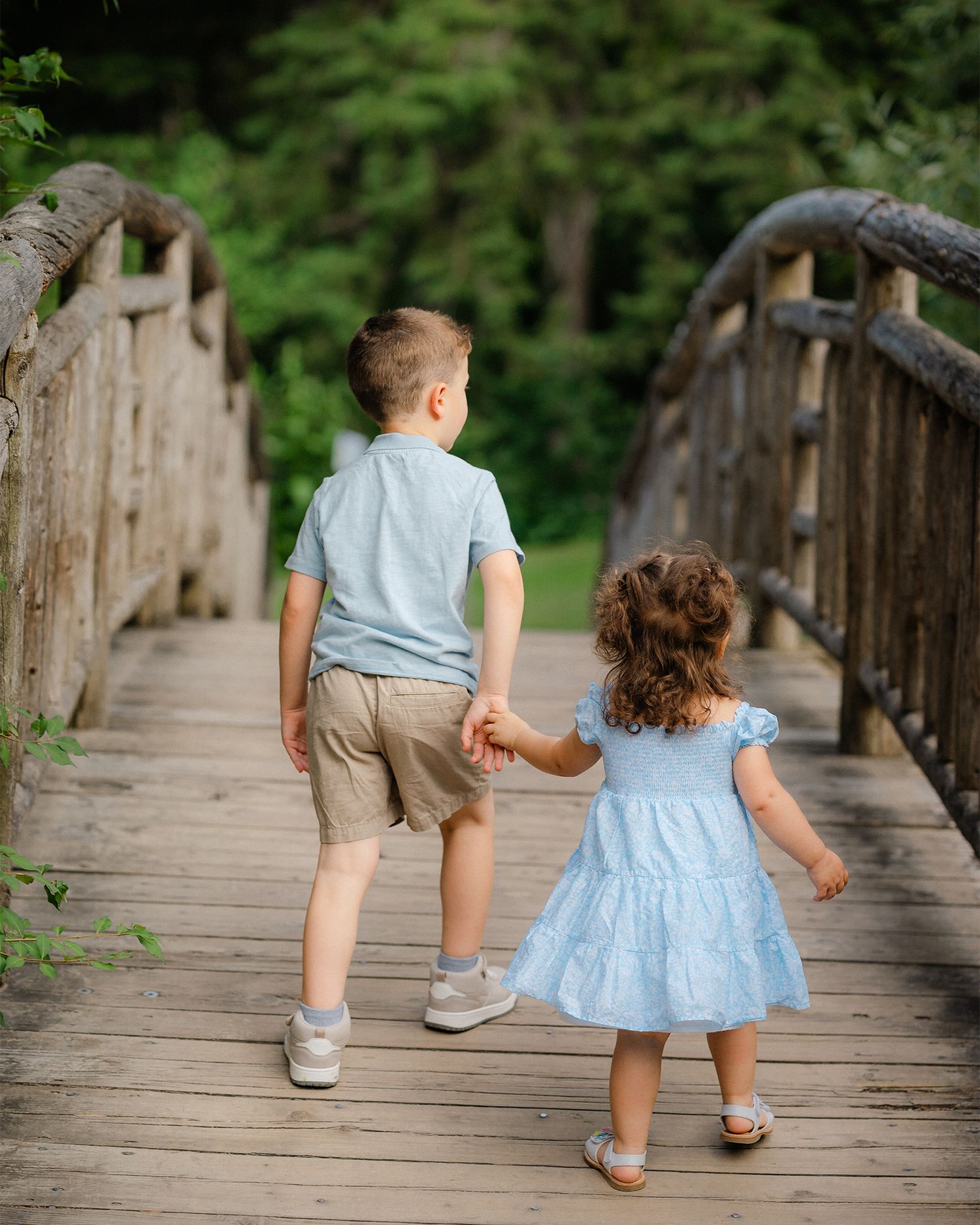 Two young children holding hands walking across a wooden bridge in a park.