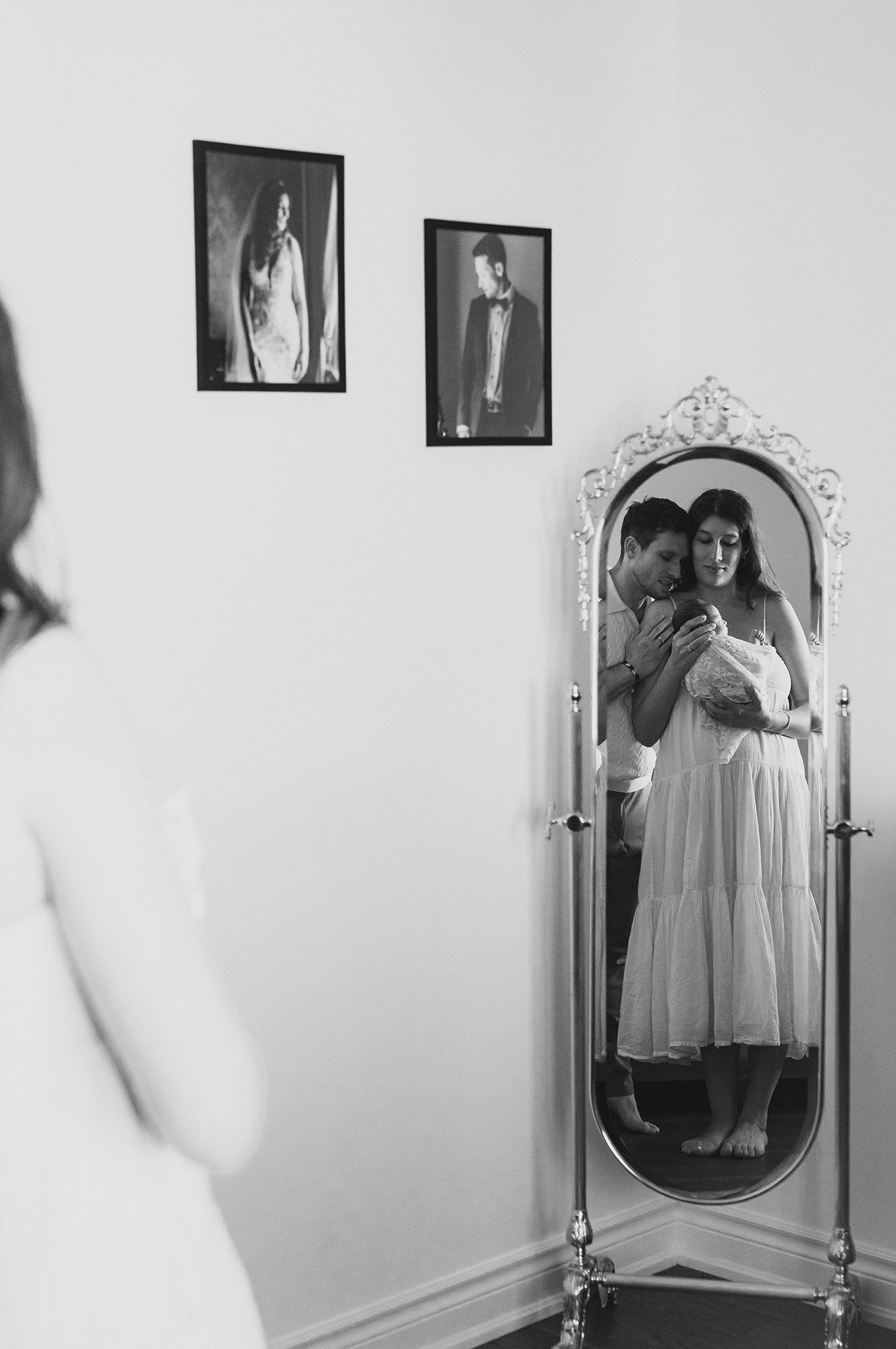 Black and white photo of a couple embracing in an ornate standing mirror, the woman in a white dress holding a newborn baby.