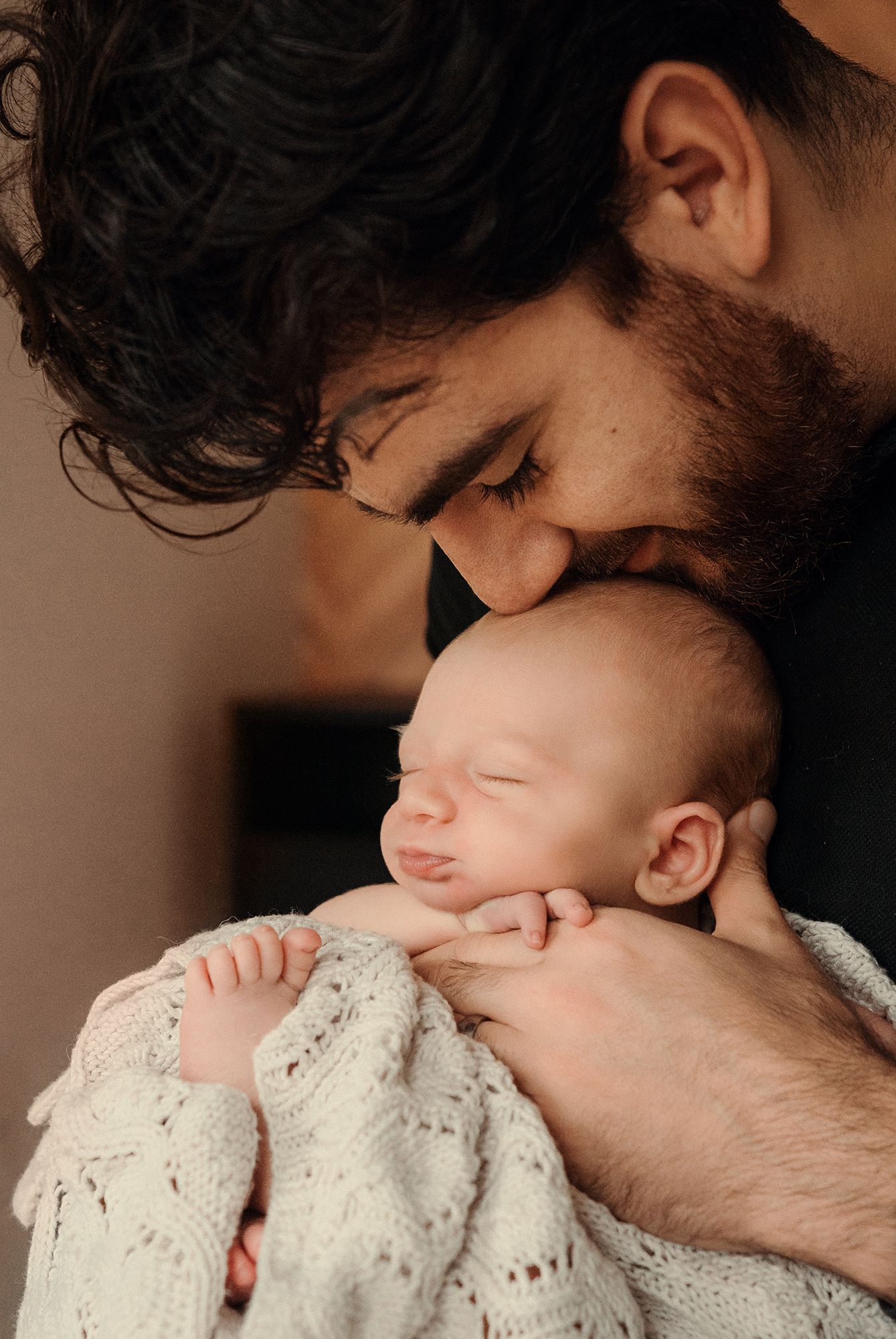 A bearded father gently kisses his sleeping newborn baby wrapped in a white knit blanket.