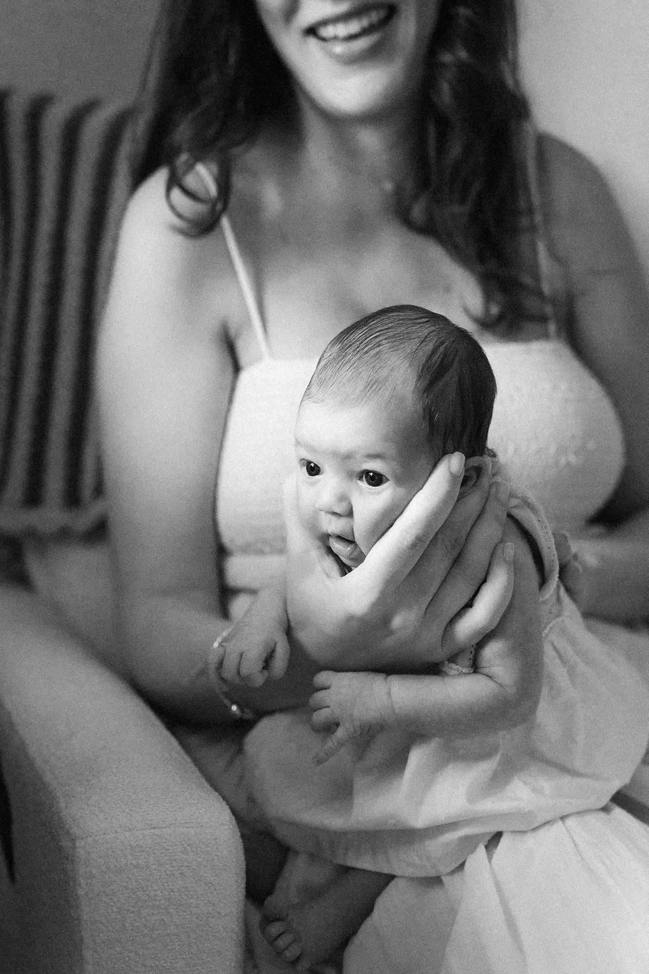Black and white photo of a smiling mother in a sundress gently holding her wide-eyed newborn baby against her chest.