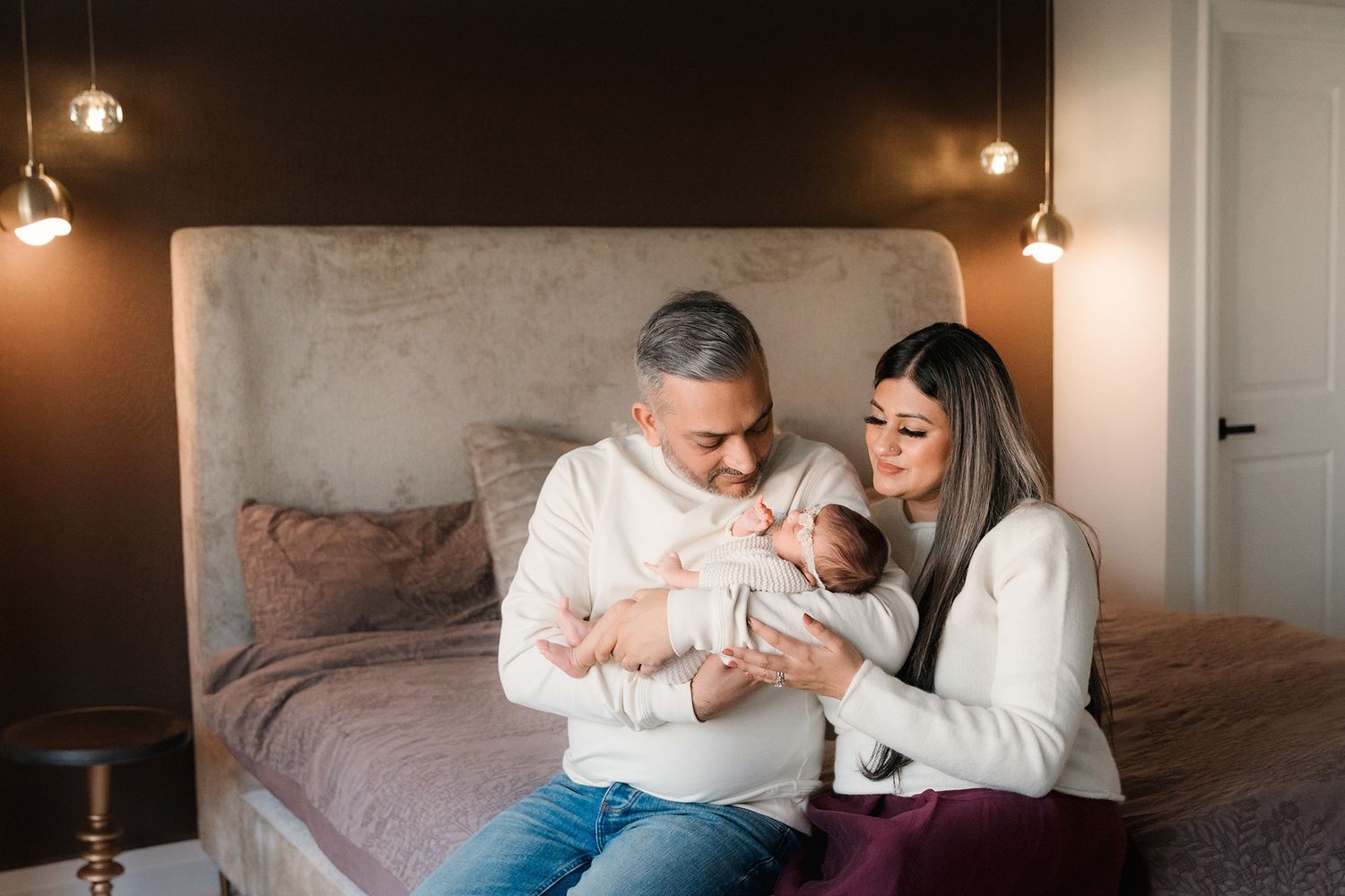 Happy parents sit together on a bed holding their newborn baby in a warmly lit bedroom.