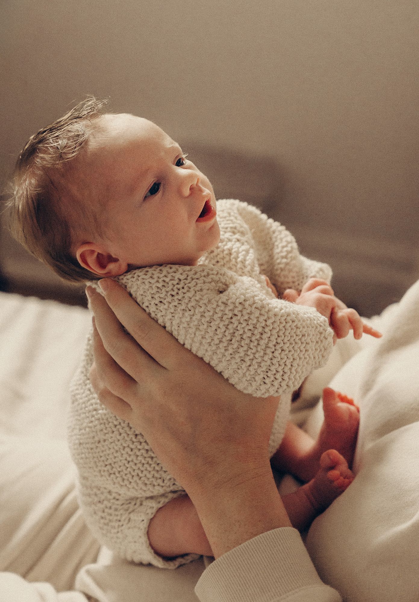 Alert newborn baby in cream knitted sweater being gently held up by parents hands on white bed.