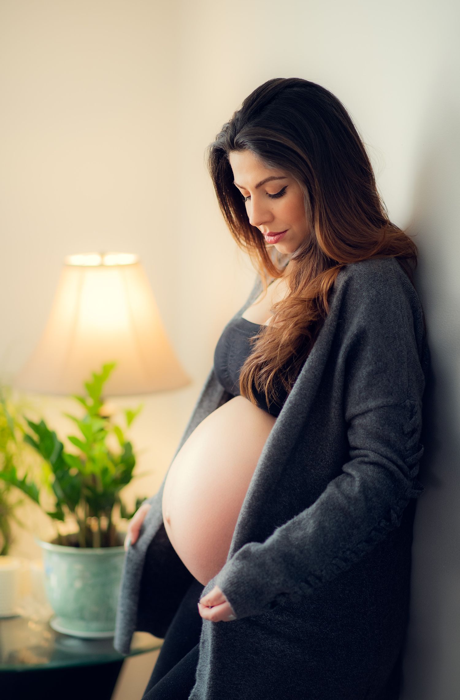 Pregnant woman in gray cardigan cradling baby bump while standing by lamp indoors.